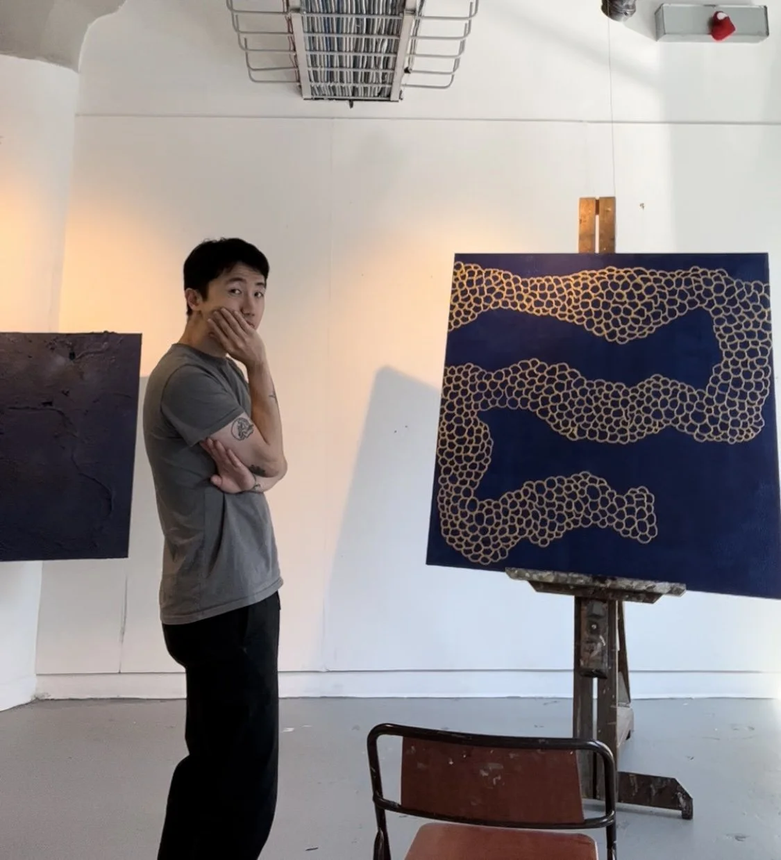 A person standing in an art studio, looking curiously at a large abstract painting on an easel. The painting features a dark blue background with a pattern of circular shapes in yellow or gold.