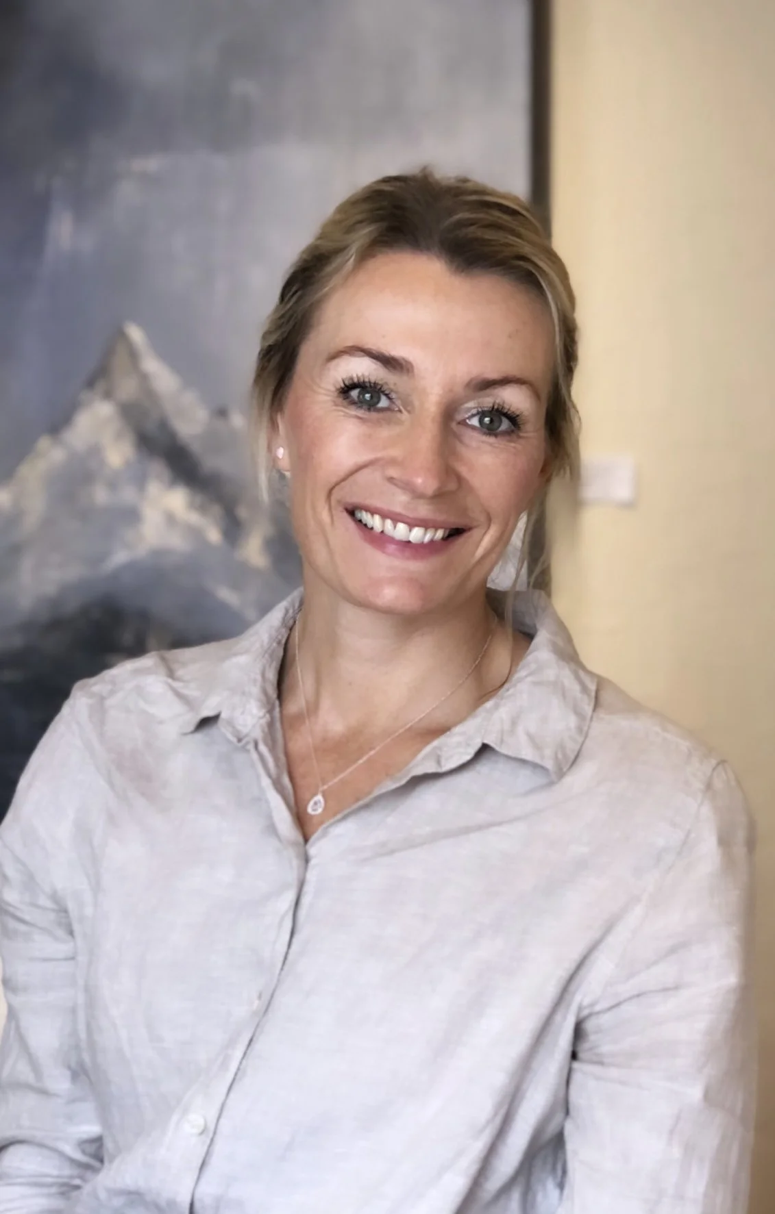 A woman smiling, wearing a light-colored collared shirt and a necklace, standing indoors with a mountain painting in the background.