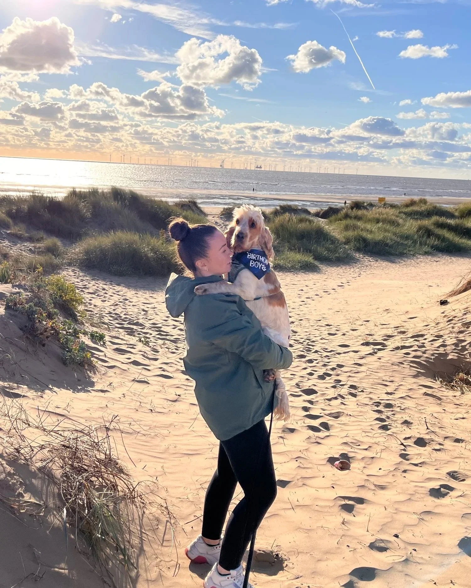 A woman holding a large dog on a sandy beach with grassy dunes, the ocean, and partly cloudy sky with wind turbines in the distance.