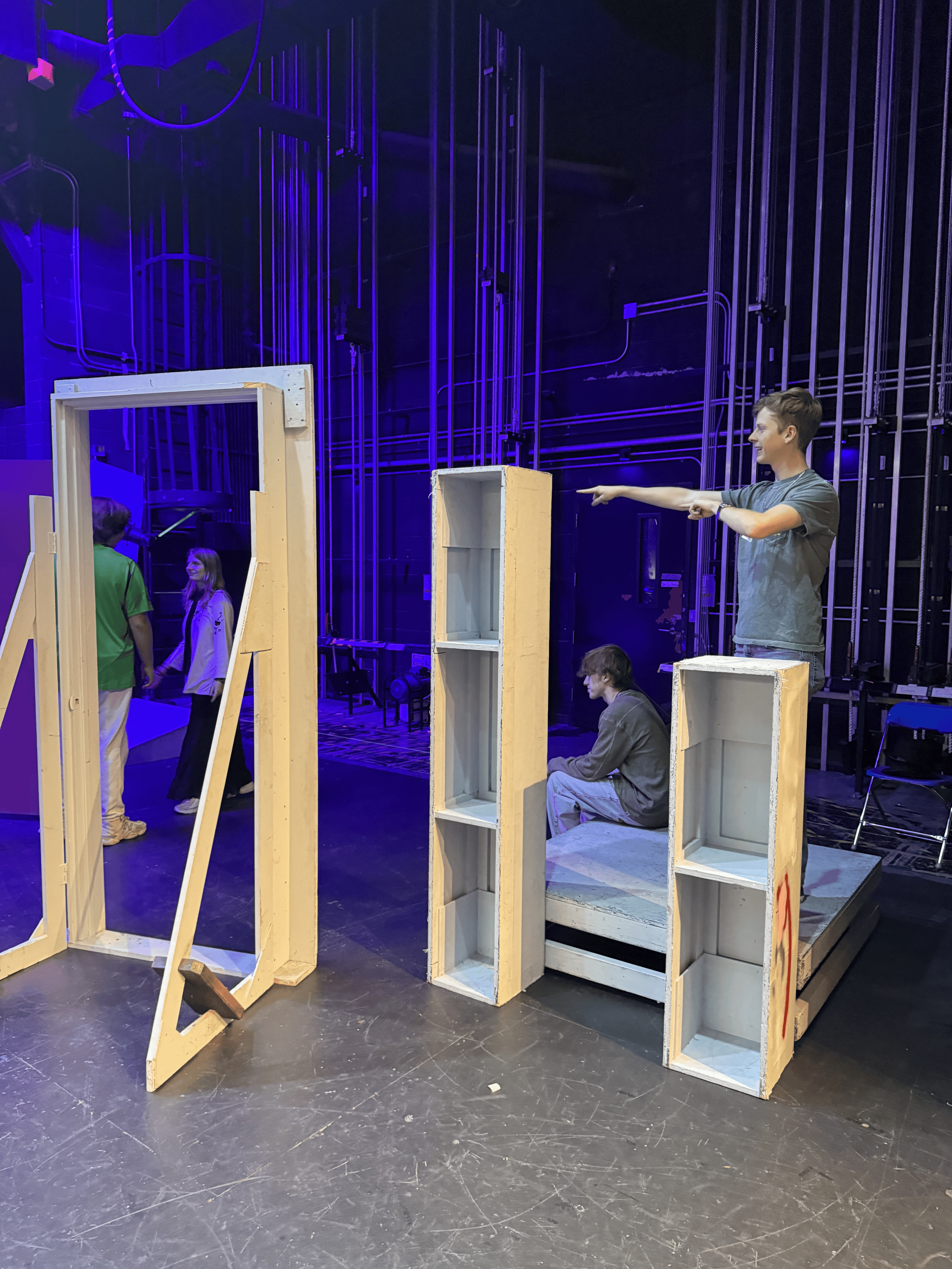 The image shows a stage set with wooden blocks and a person standing on top of one of the blocks, pointing forward, with other people in the background.