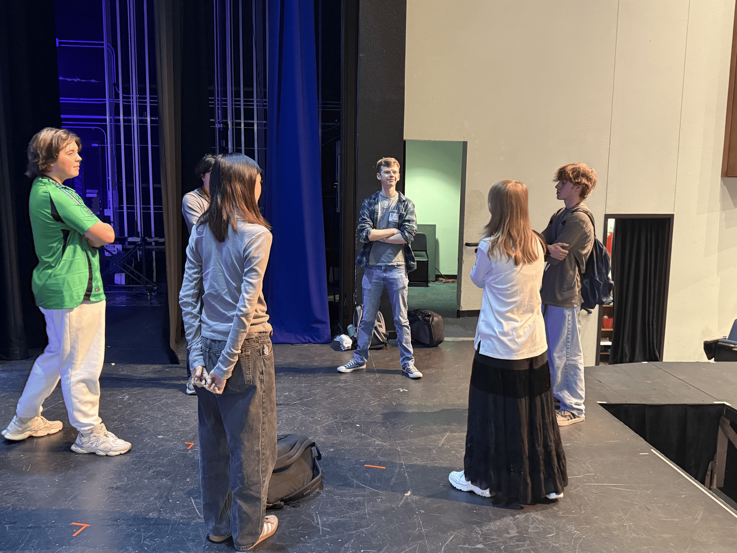 Group of seven young people standing and talking on a theater stage, some with arms crossed, near a blue curtain and a green wall, with stage equipment in the background.