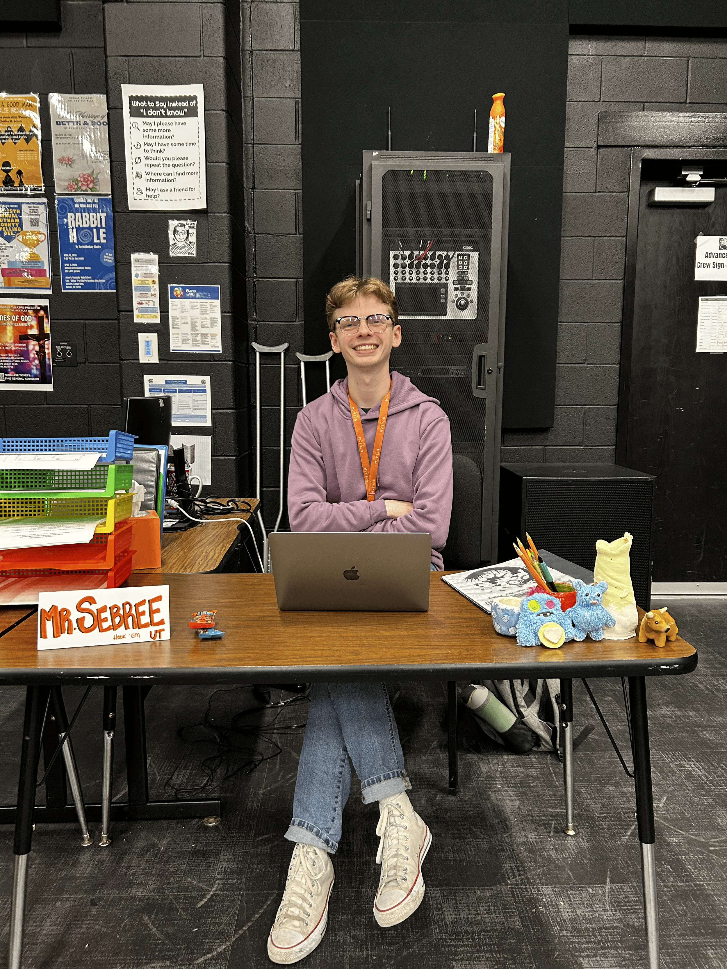 A young man with glasses and a purple hoodie sitting at a desk with a laptop. The desk has colorful craft supplies and a small plush toy. There are posters and signs on the black brick wall behind him, and a speaker and audio equipment. The sign on the desk reads 'MR. SEBREE.'