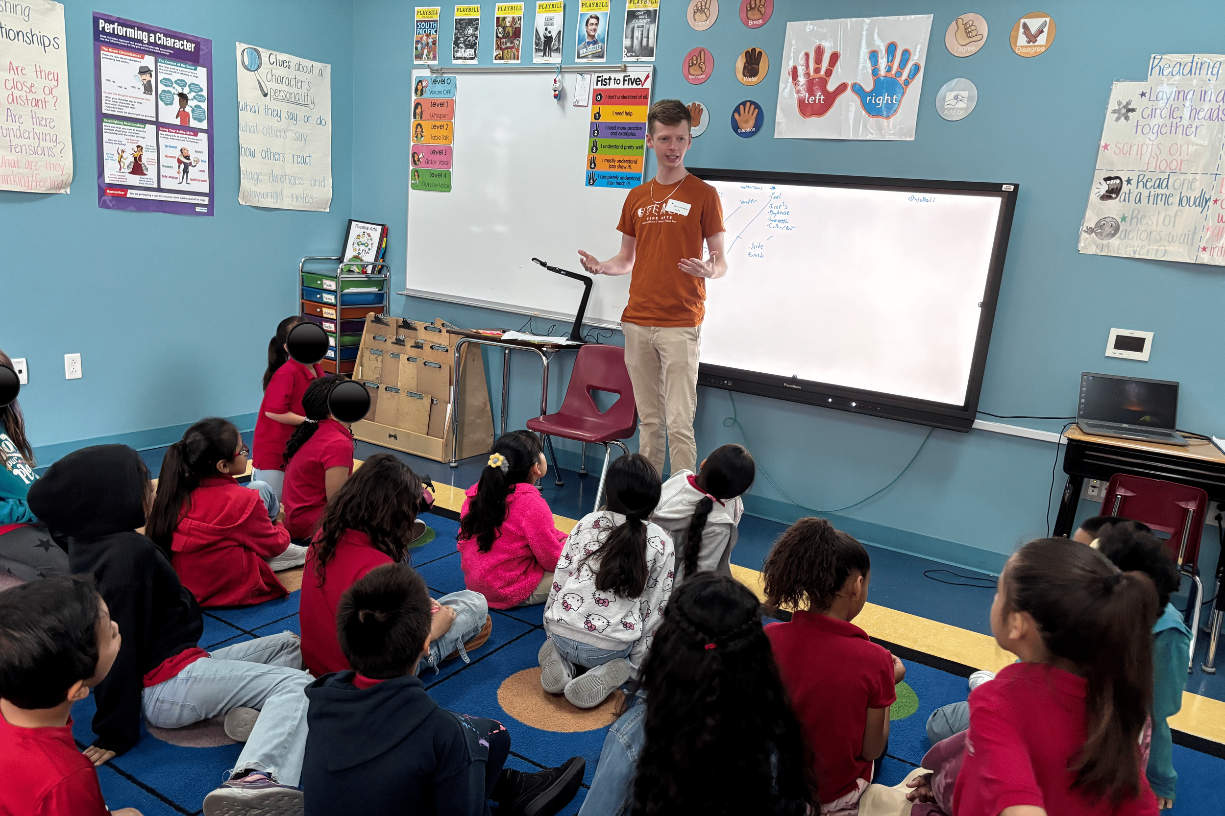 A classroom with a teacher standing at the front, giving a lesson to a group of children sitting on the carpet. The classroom has colorful posters, a whiteboard, and educational materials on the walls.