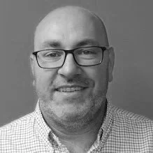 A black and white photo of a man with glasses, a beard, and a checkered shirt, smiling.