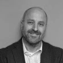 Black and white photo of a man with a beard, wearing a collared shirt and blazer, smiling at the camera.