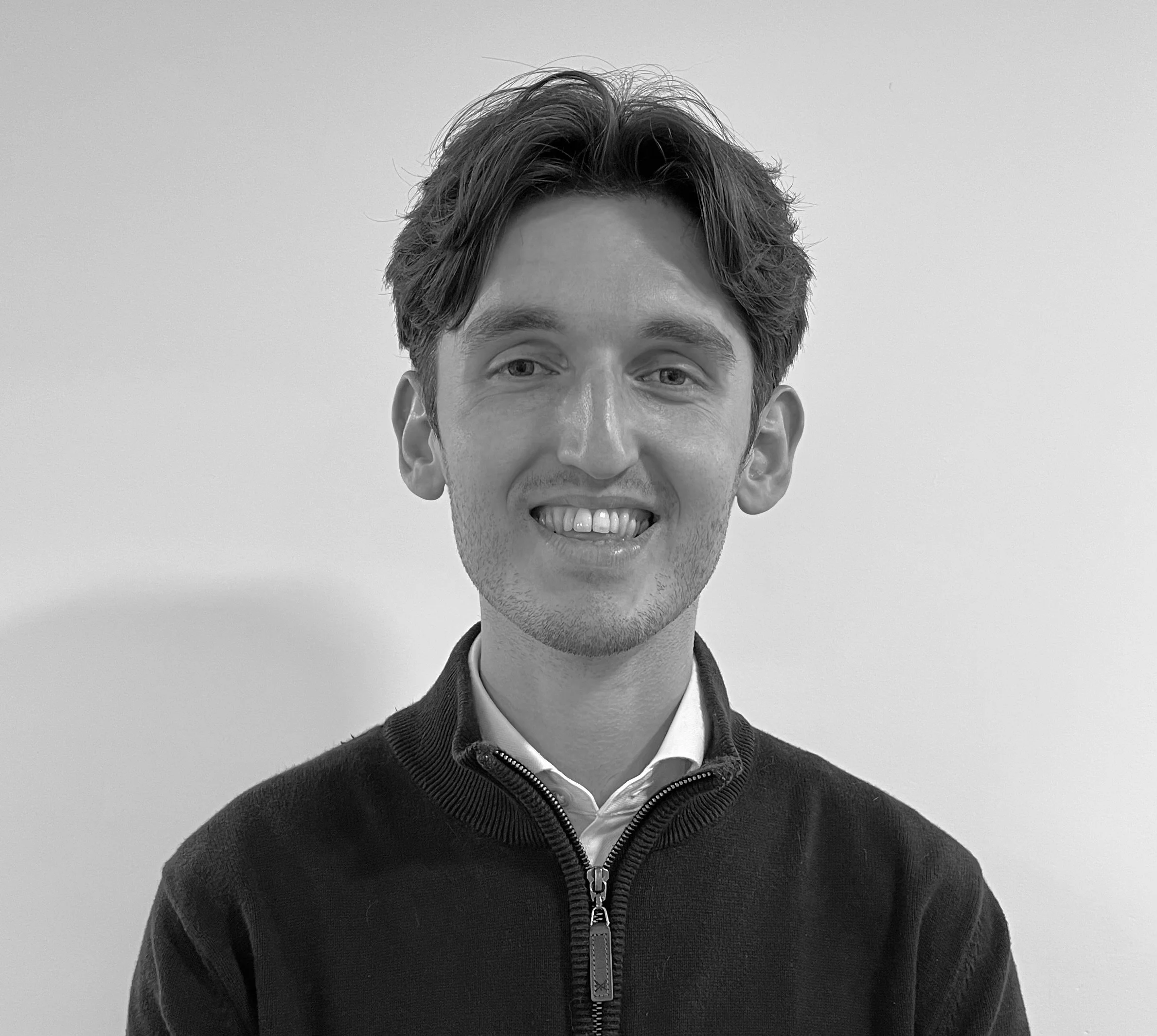 Black and white portrait of a young man smiling, wearing a zip-up sweater over a collared shirt, against a plain background.