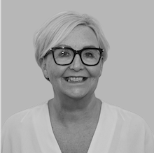 A smiling woman with short blonde hair and glasses, wearing a light-colored top.