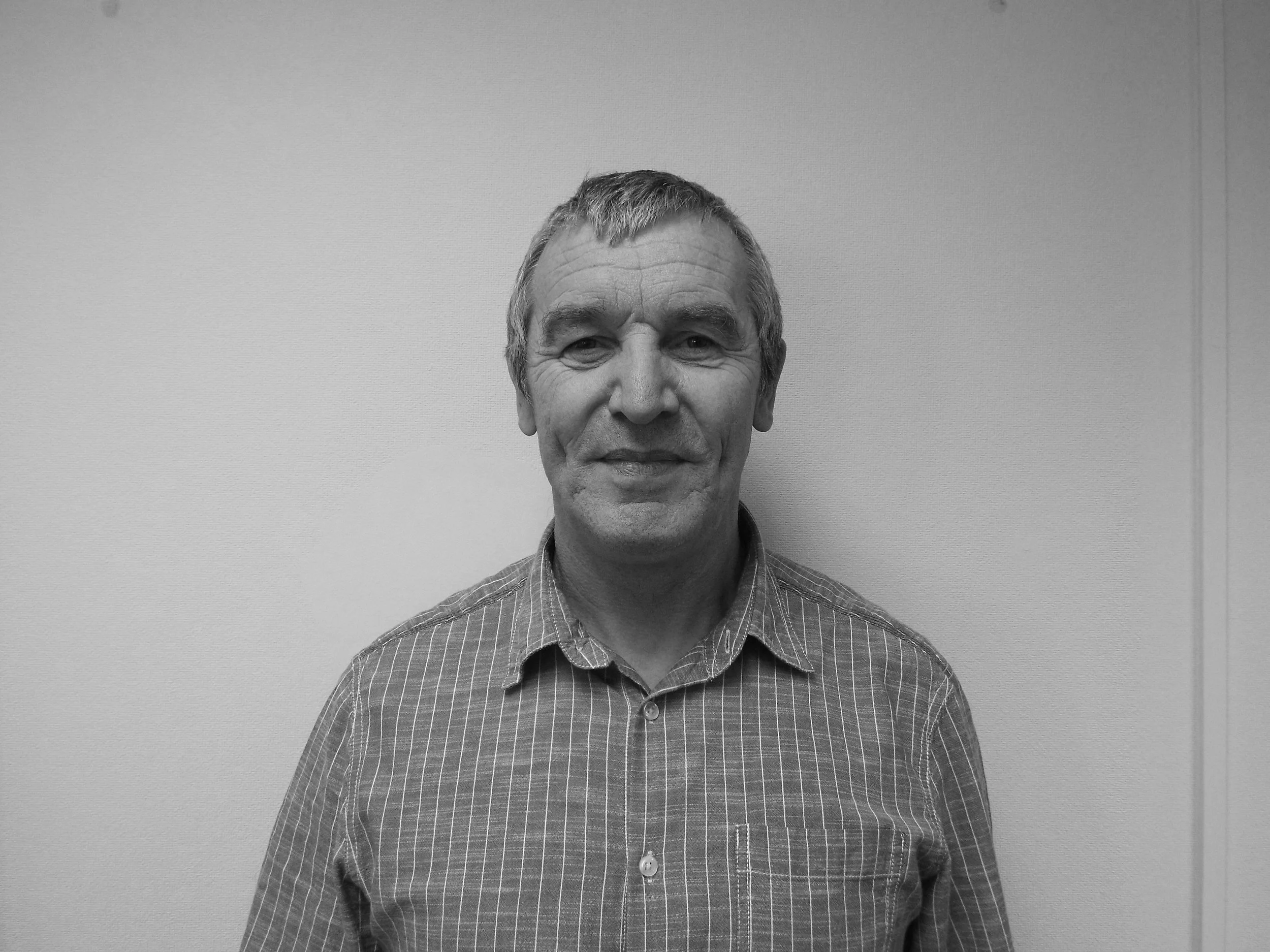 Black and white photograph of a smiling middle-aged man with short hair, wearing a checkered button-up shirt, standing against a plain wall.