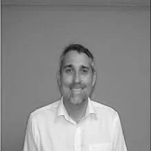 A man with short dark hair and a beard, smiling and wearing a white dress shirt, standing against a plain wall.