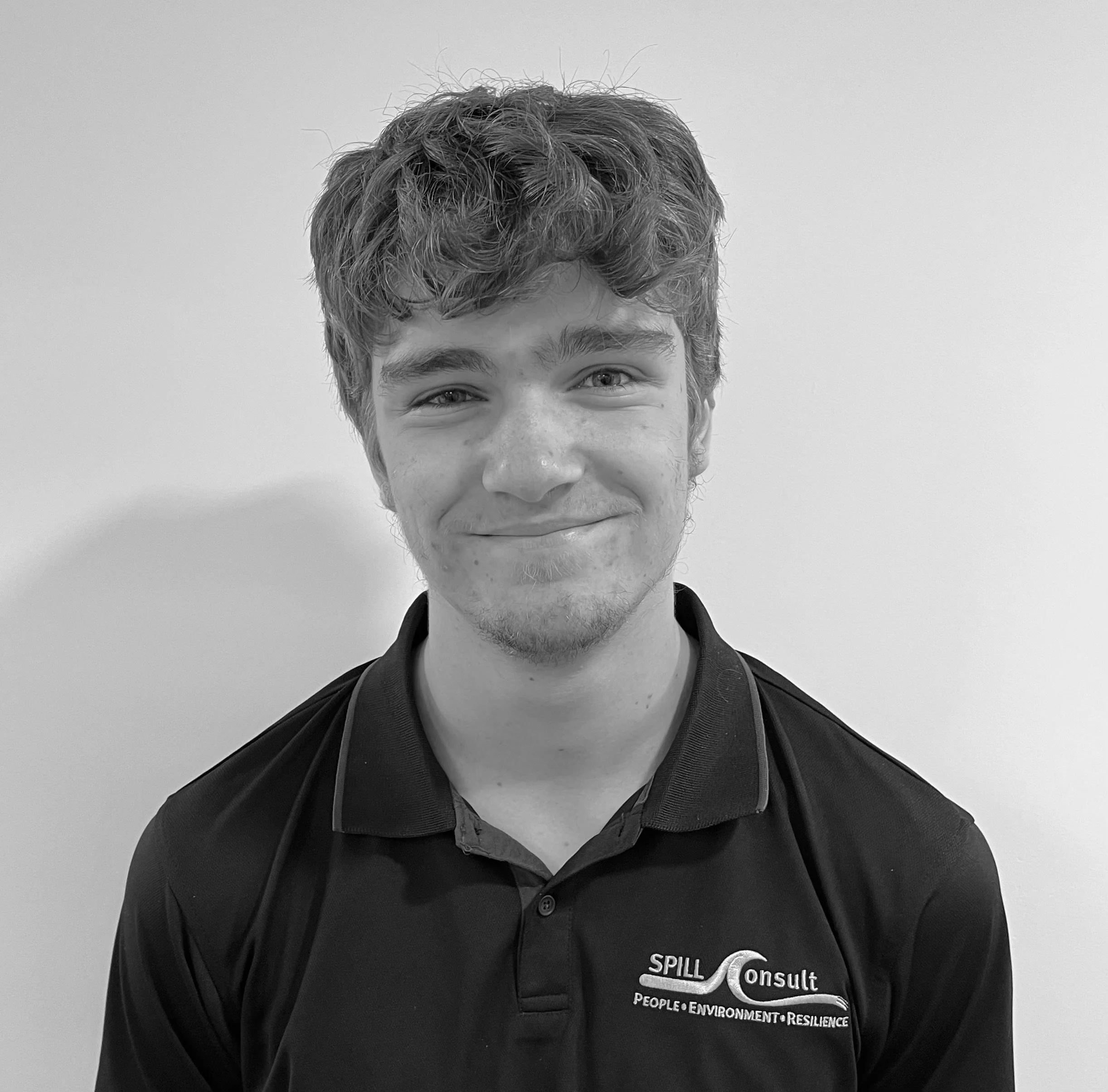 Young man with wavy hair wearing a black polo shirt with the Spill Consult logo, smiling against a plain background.