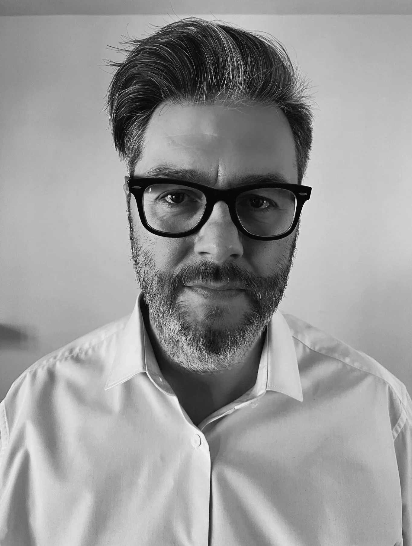 Black and white portrait of a man with glasses, a beard, and styled hair wearing a collared shirt.