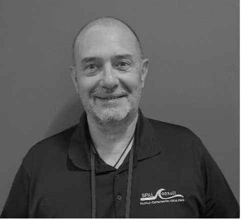 Black and white photo of a smiling man with a beard, wearing a collared shirt and a branded jacket, standing against a plain background.