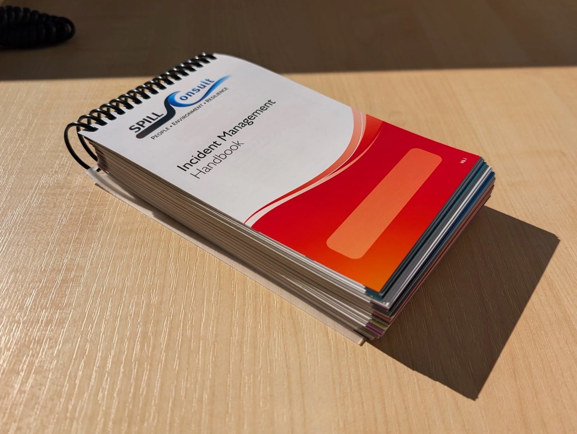A spiral-bound handbook titled "Spill Consulting: People and Environment Resilience, Incident Management Handbook" on a wooden desk.