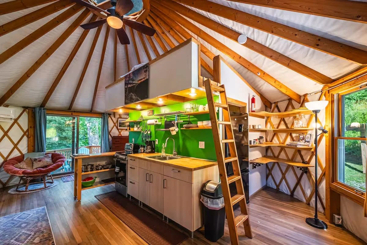Interior of the cozy, unique yurt with a small kitchen area featuring green backsplash, wooden shelves, and a ladder leading to a loft in New River Gorge WV