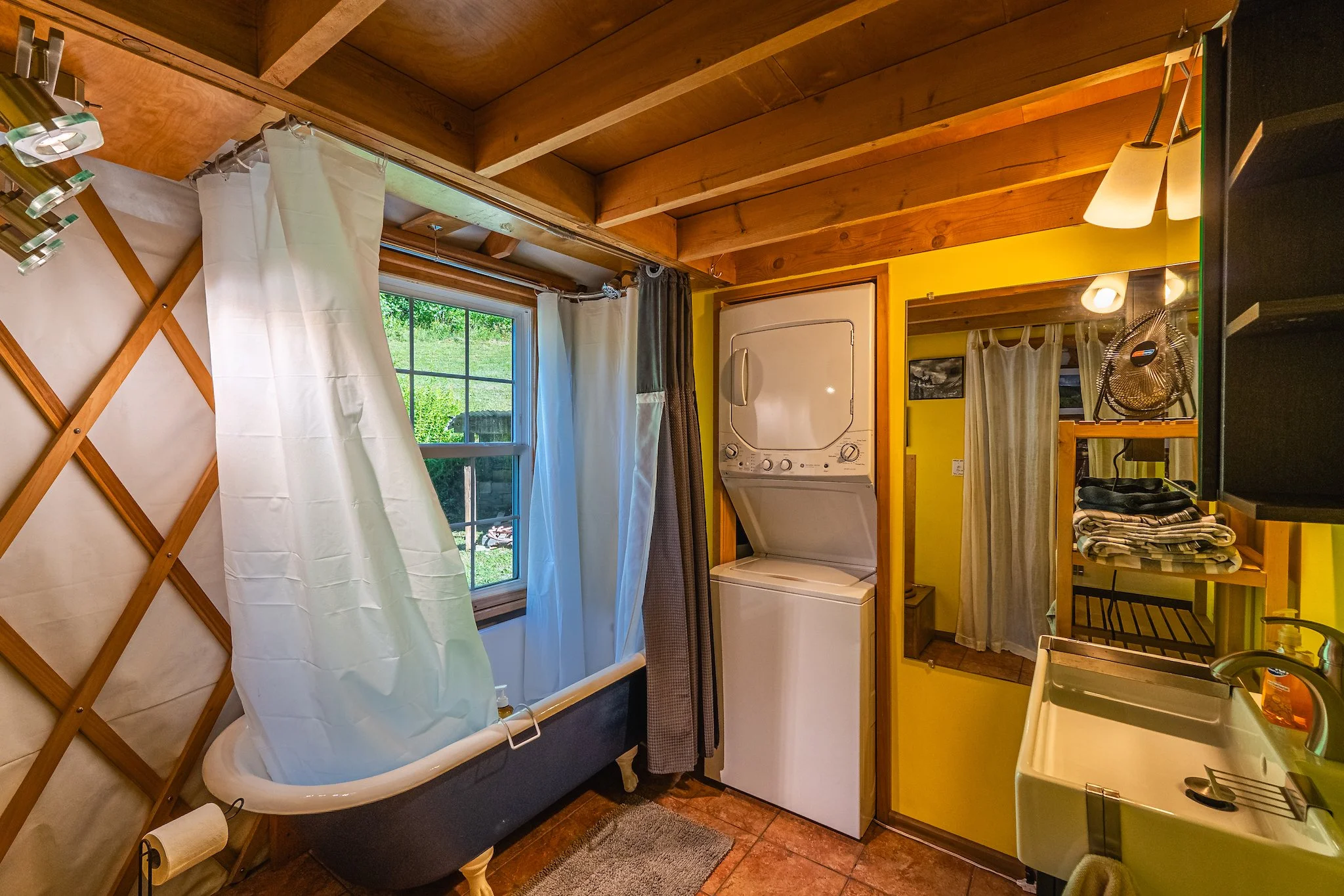 Rustic bathroom in the cozy yurt at New River Nooks in Fayetteville WV with a clawfoot bathtub, washer and dryer, and shelves with folded towels.