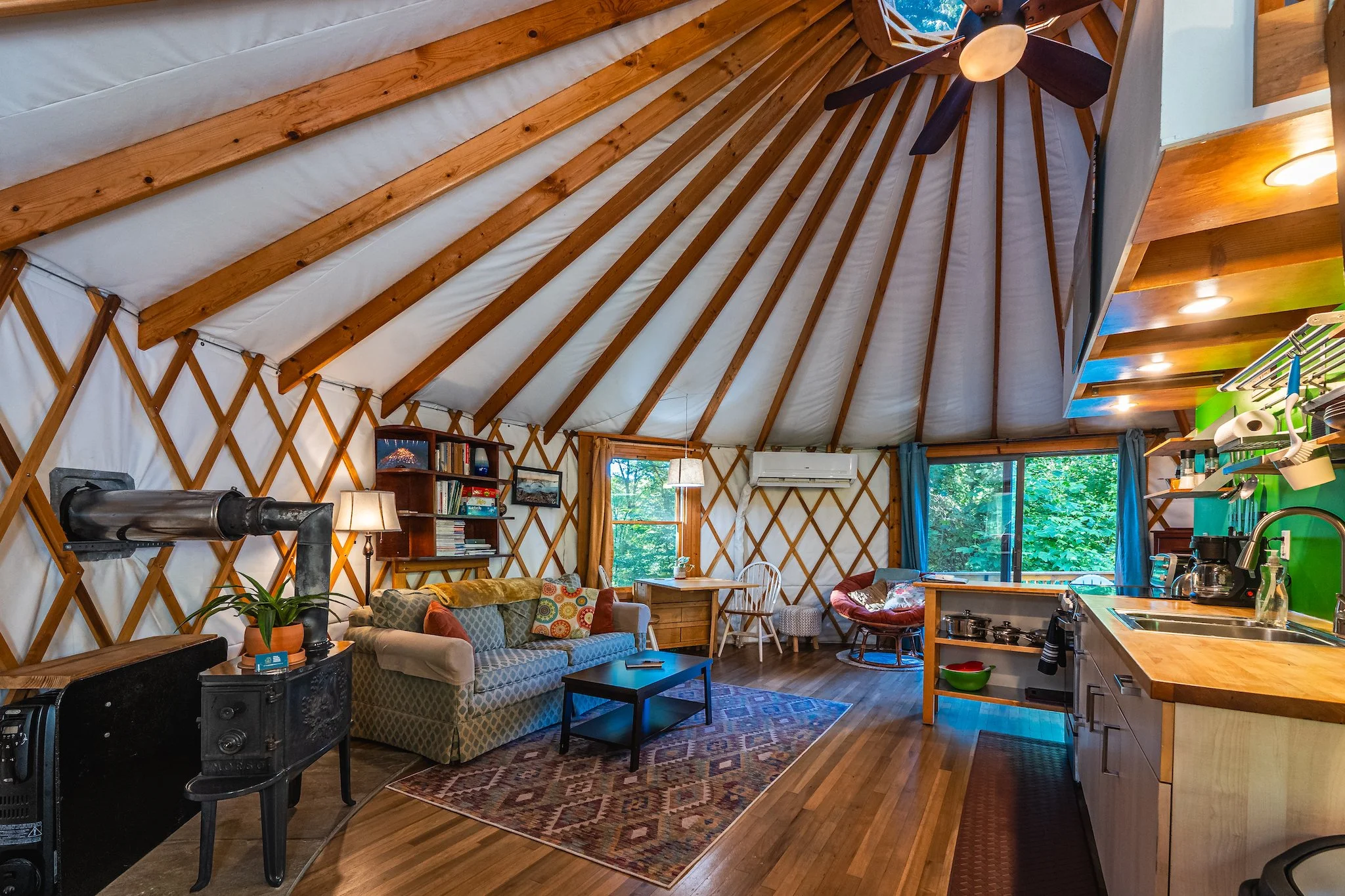 Interior of the yurt vacation rental with a living area with a couch, armchair, and bookshelf, a dining table near the windows, and an open kitchen with a sink and appliances in New River Gorge WV.