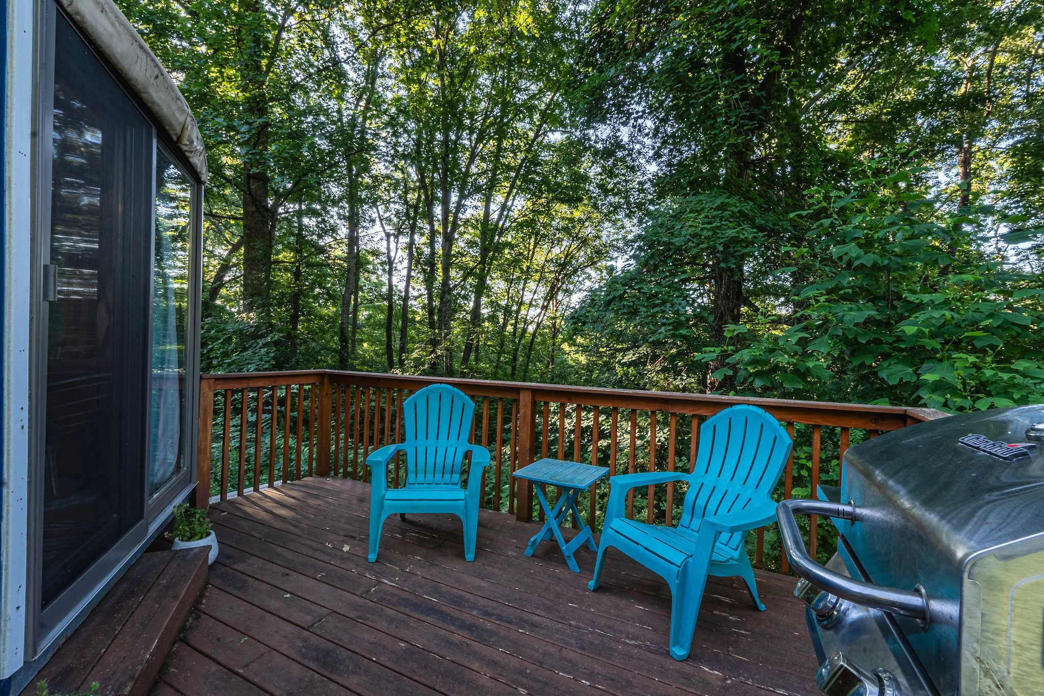 A wooden deck off of the unique yurt with two blue Adirondack chairs and a small blue table surrounded by green trees in New River Gorge WV.