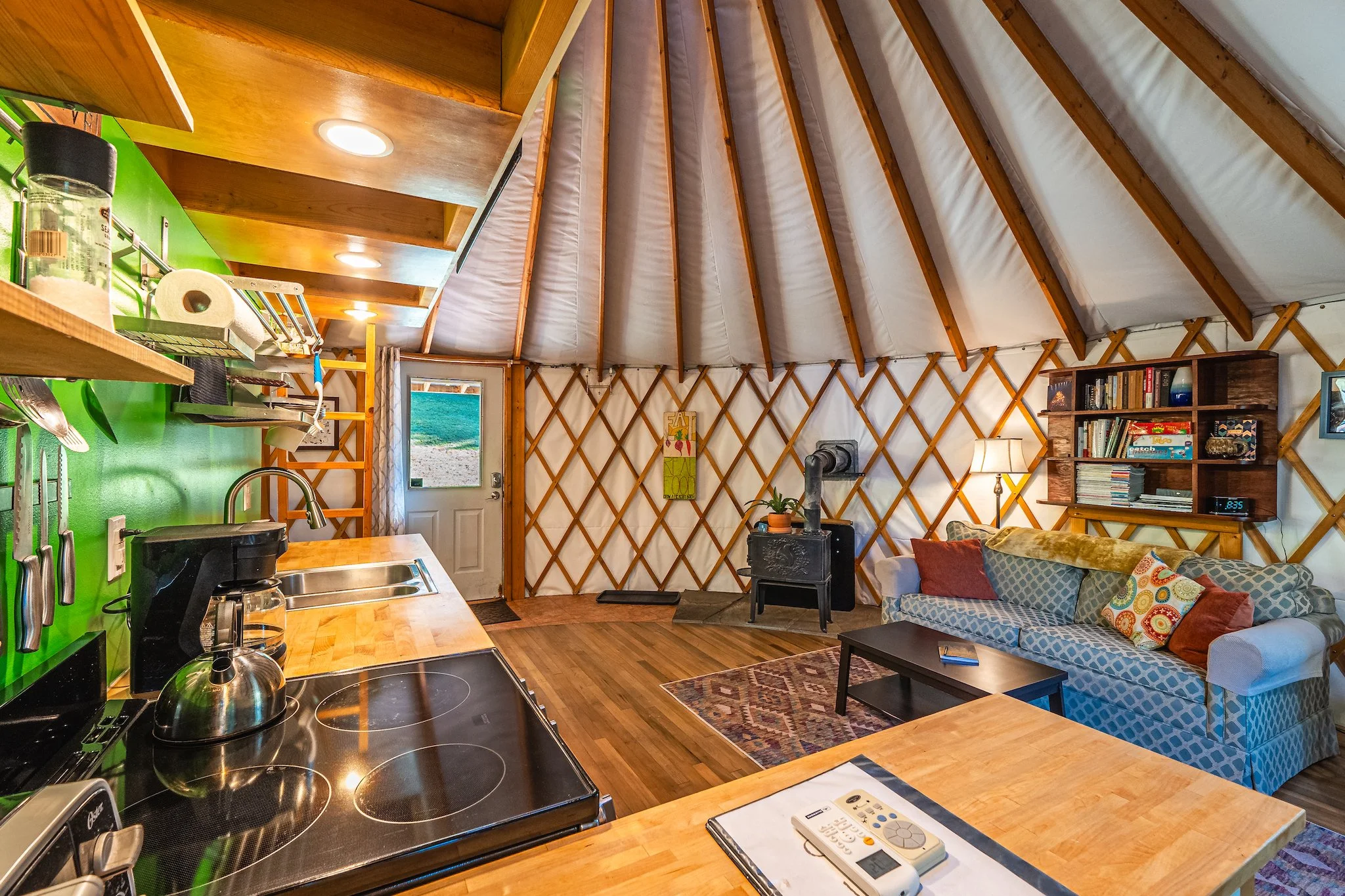 Interior of a yurt at New River Nooks with cozy living space with a wood stove, a bookshelf, a patterned rug, a couch with colorful pillows, and a kitchen counter in New River Gorge WV.