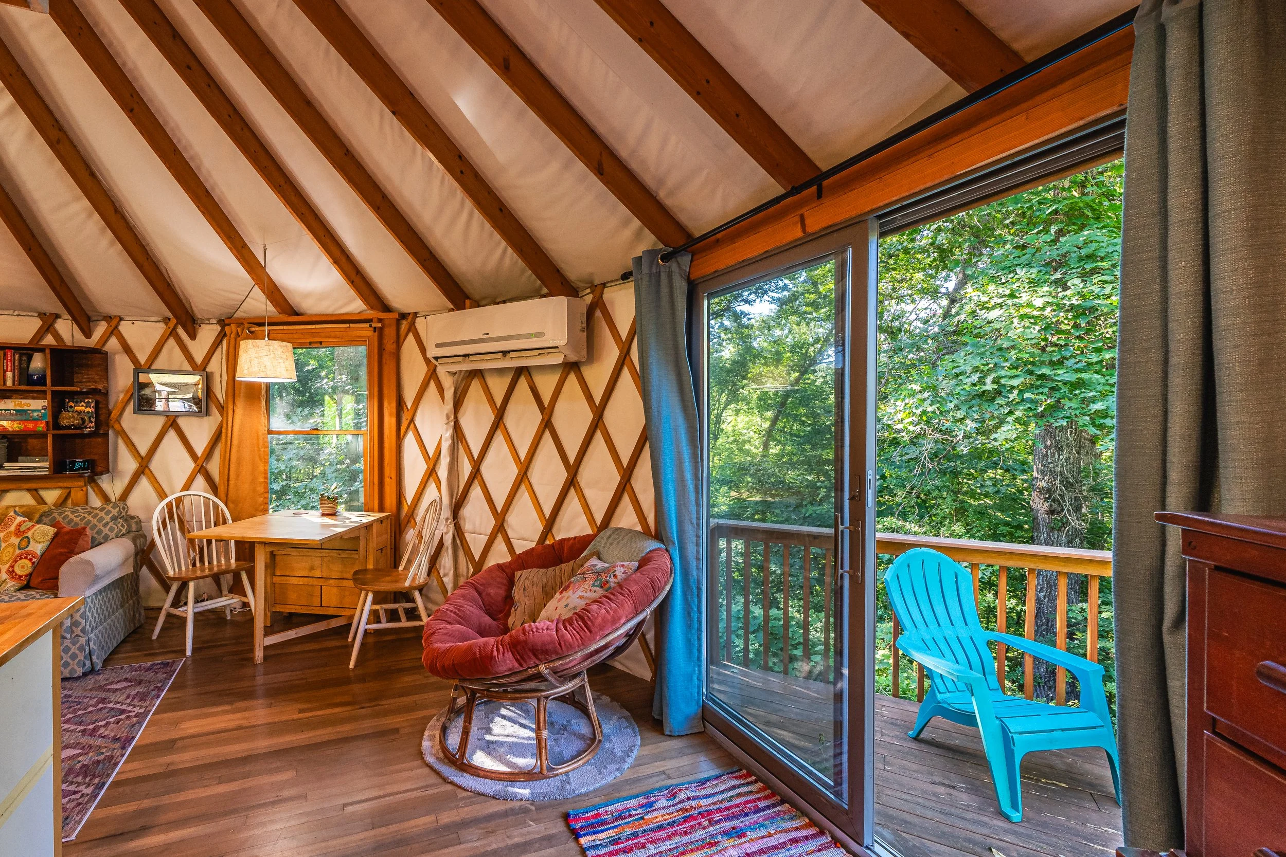 Interior of a cozy yurt with a sliding glass door leading to the deck with a blue Adirondack chair, surrounded by lush green trees in New River Gorge WV.