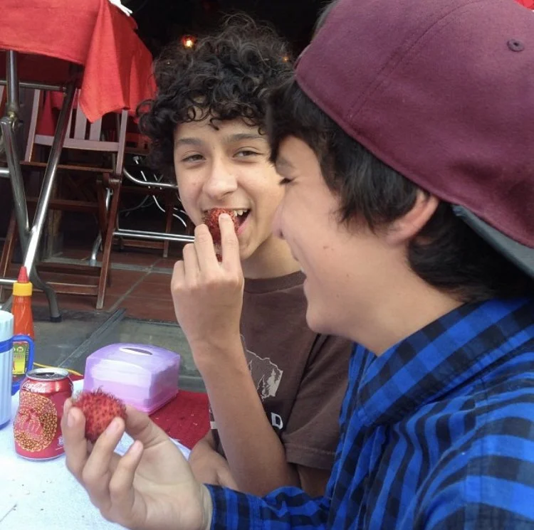 Two teenage boys laughing together as they are eating fruit.