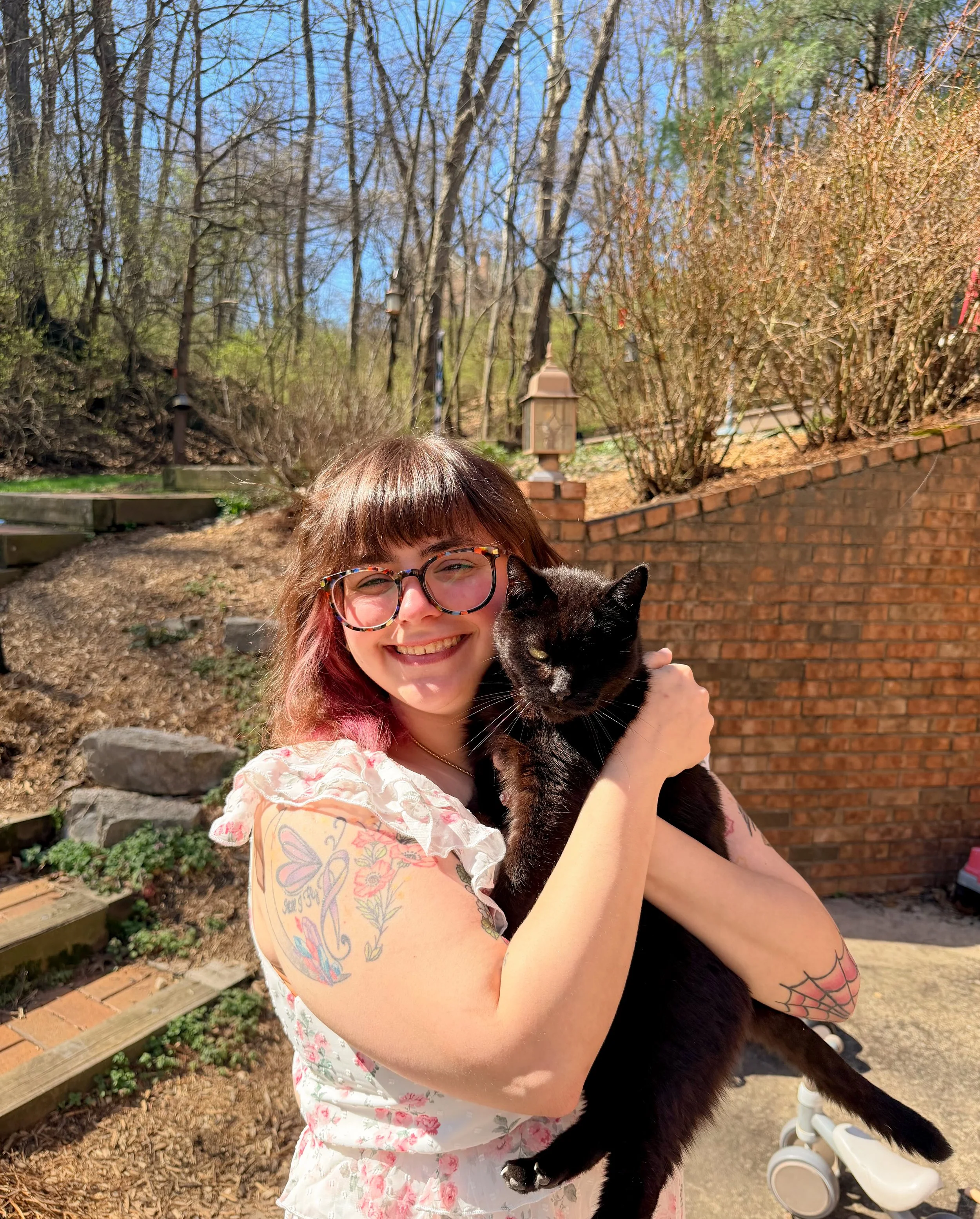 A smiling woman with glasses and floral tattoos on her arms holding a black cat outdoors on a sunny day, with trees and a brick wall in the background.