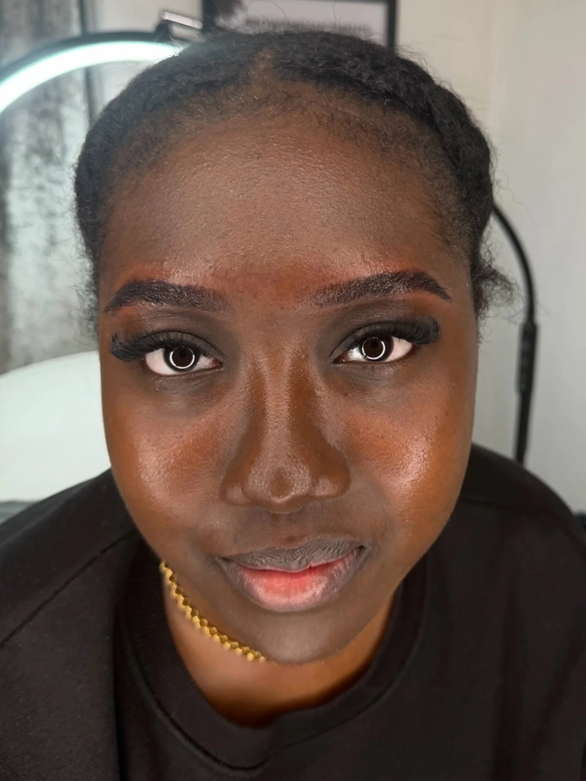Close-up of a woman with smooth dark skin, styled hair, bold eye makeup, and a gold necklace, looking directly at the camera.