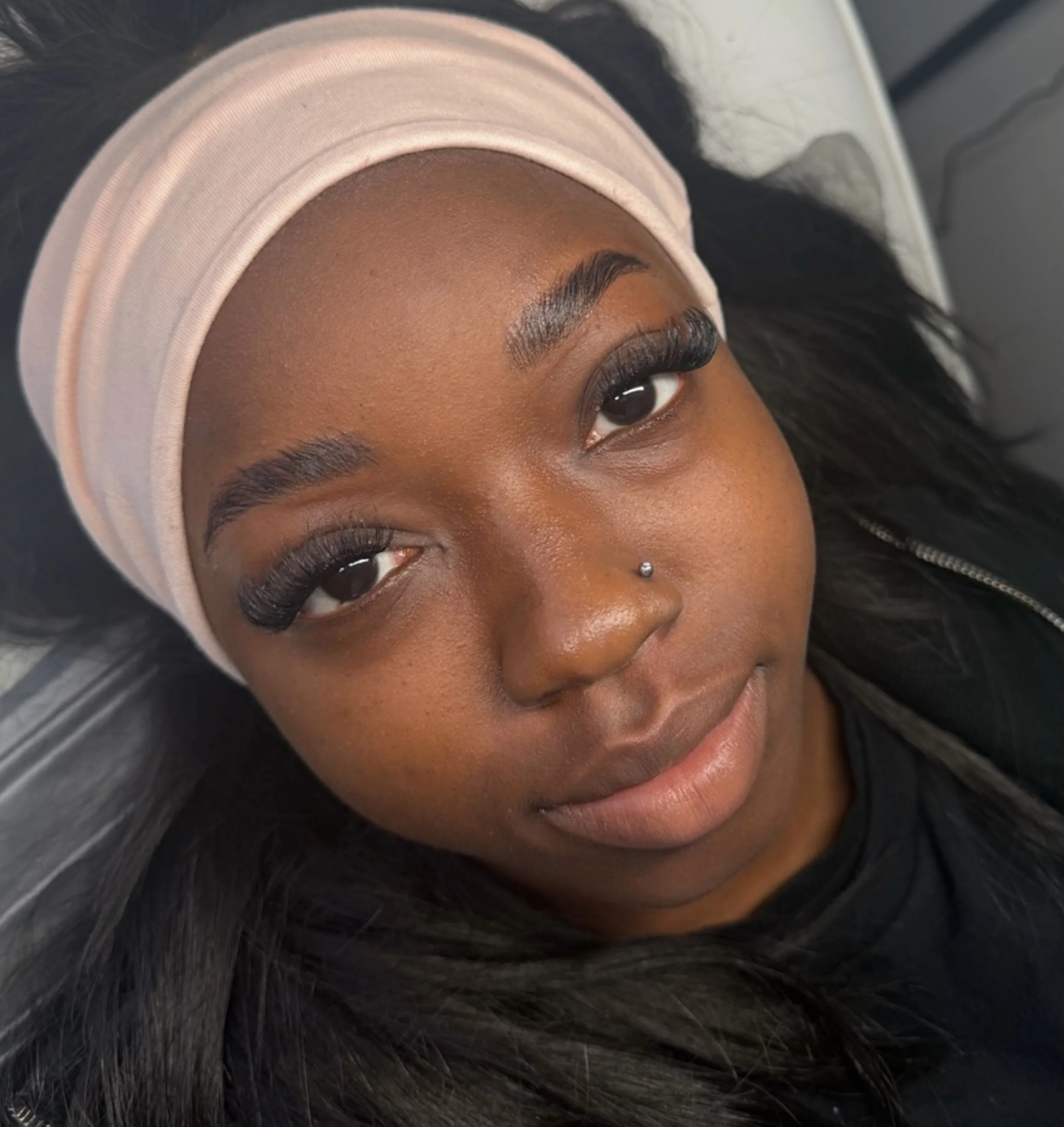 A young woman with dark hair, wearing a light pink headband and black clothing, looking at the camera with makeup and long, curled eyelashes.