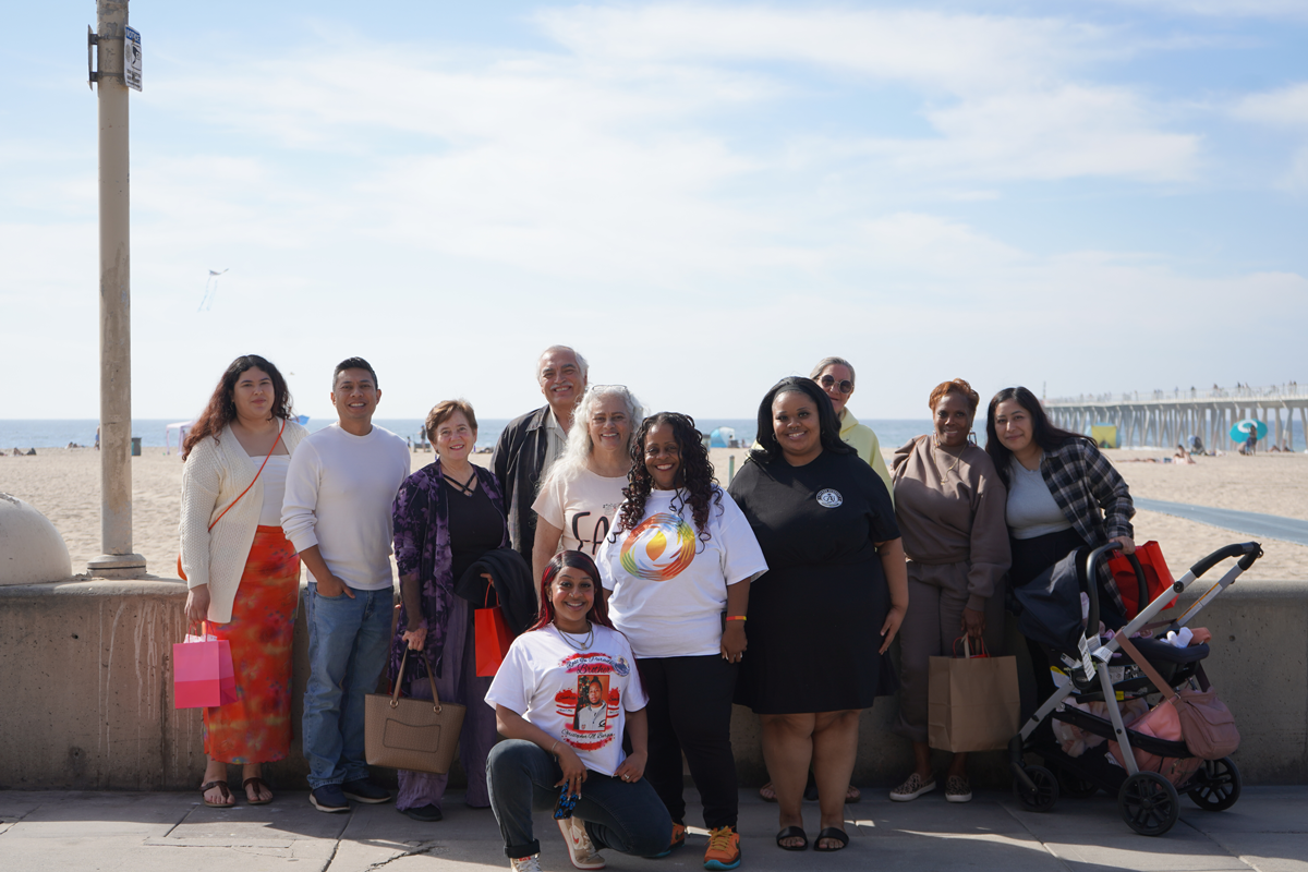 Group of ten women and one young girl standing on a beach promenade, smiling, with ocean and pier in the background. Some hold bags, and one has a stroller.