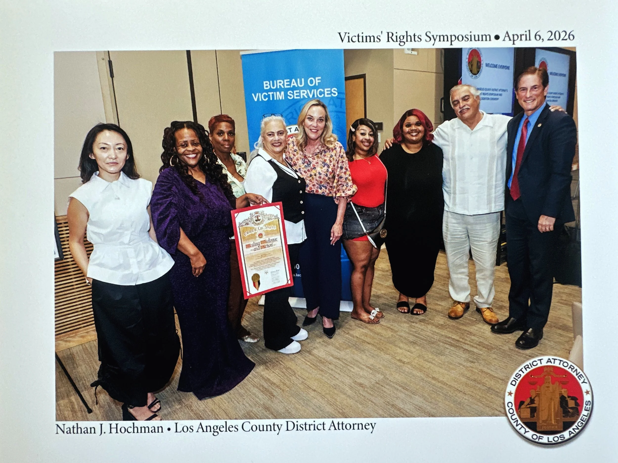 Group of people standing together at the Victims' Rights Symposium on April 6, 2026, in front of a blue 'Bureau of Victim Services' banner, with some holding documents or certificates.