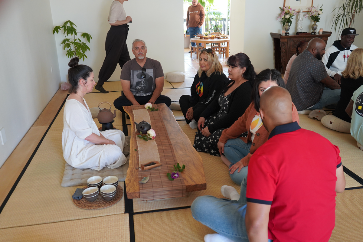 People participating in a traditional tea ceremony indoors, sitting on tatami mats around a low wooden table with teapots, tea cups, and flowers, while a woman in white leads the ceremony.