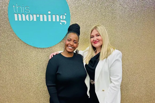 Two women smiling and standing close together in front of a wall with a blue circle that says "this morning." One woman has dark skin and black hair in a bun, wearing a black sweater. The other has fair skin, blonde hair, and is wearing a white blazer over a black top.
