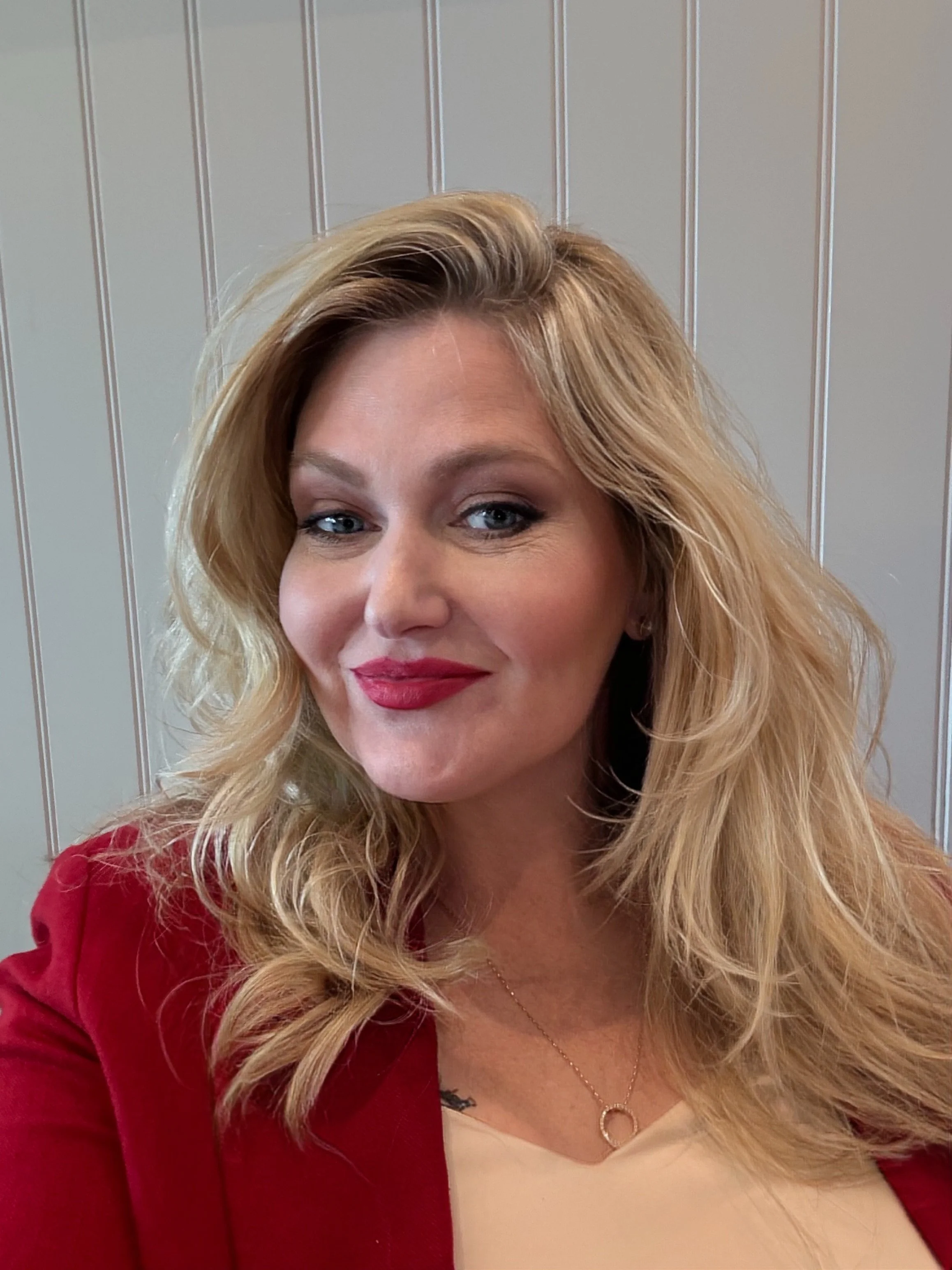 A woman with blonde, wavy hair, blue eyes, red lipstick, wearing a beige top and a red jacket, sitting against a wall with vertical paneling.