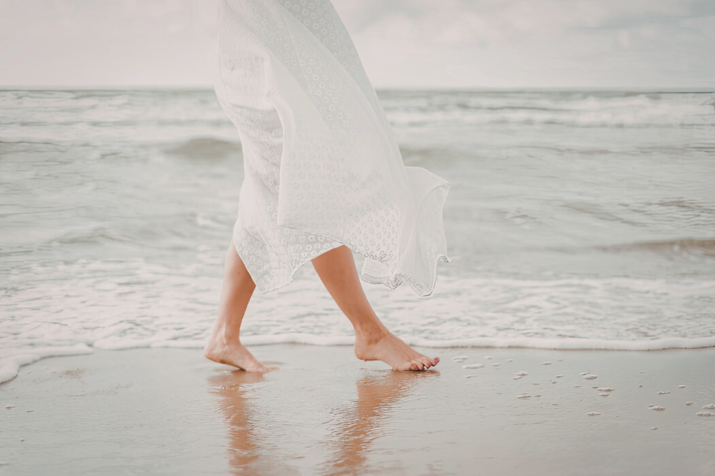 Person walking barefoot on the beach, wearing a white dress, with gentle waves in the background.