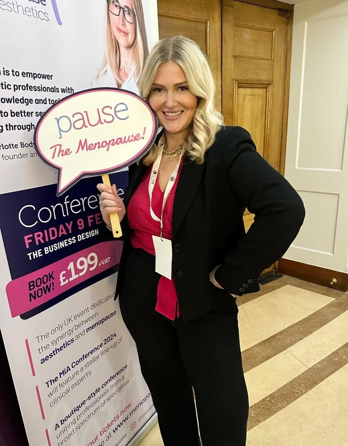 A woman with blonde hair wearing a black blazer and pink top is smiling and holding a speech bubble sign that says "pause The Menopause!" She is standing indoors next to a conference banner about a menopause event.