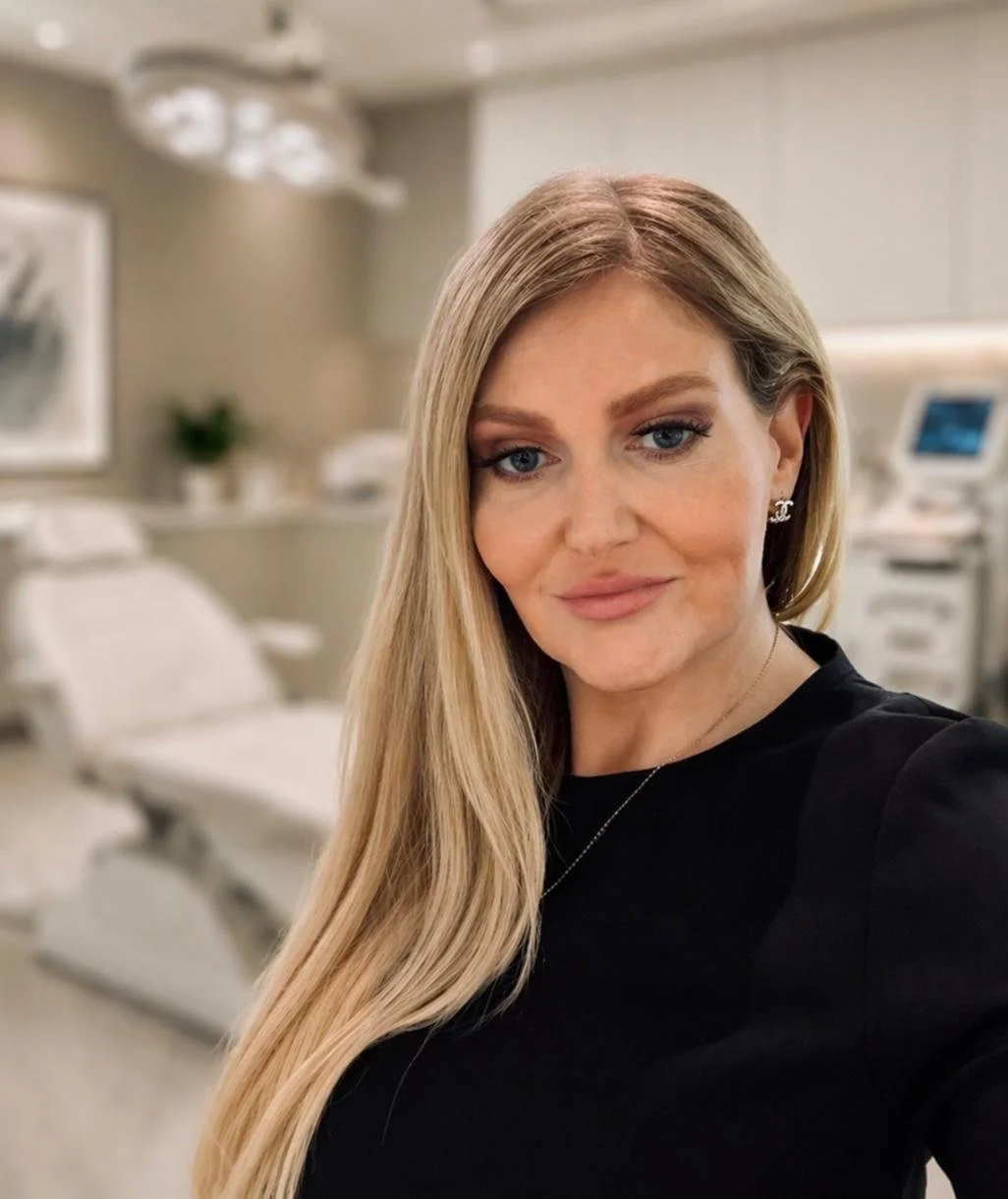 A woman with long blonde hair taking a selfie in a medical examination room.
