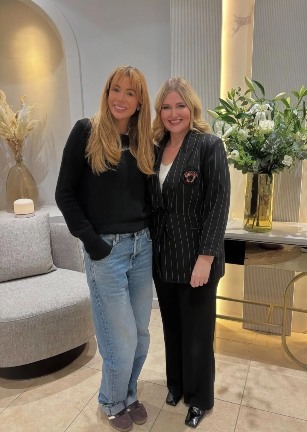 Two women standing side by side, smiling indoor setting with a large floral arrangement and modern decor in the background.