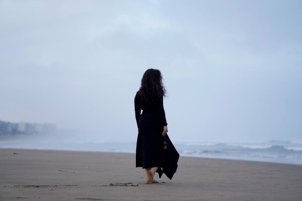A woman with long curly hair wearing black clothing walks barefoot on a deserted beach, holding a black bag with a red rose. The ocean waves are visible, and the sky is overcast.