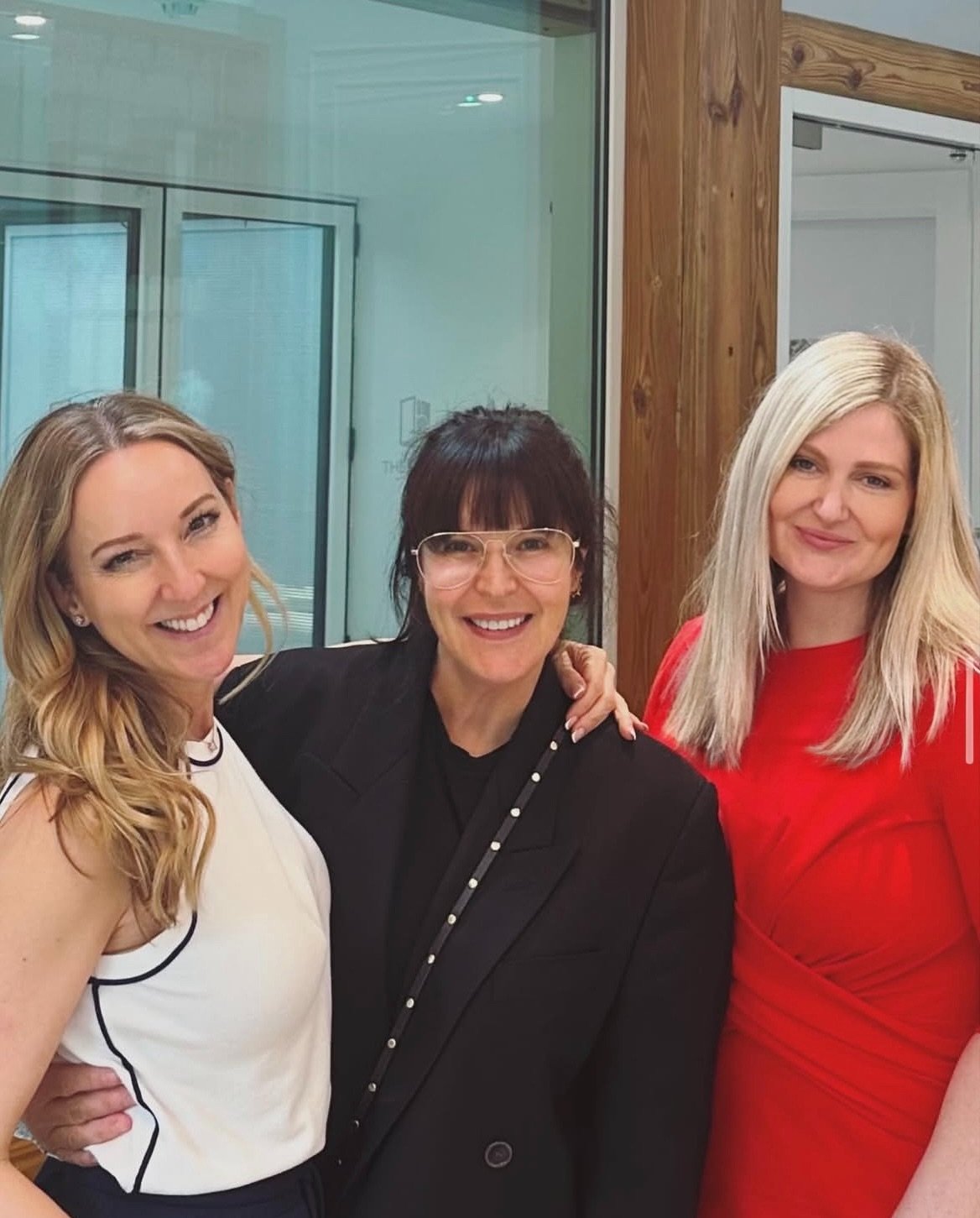 Three women smiling and posing together indoors. One woman is wearing a white top, the second woman is in a black blazer with glasses, and the third woman is in a red dress. They are standing close together with arms around each other.