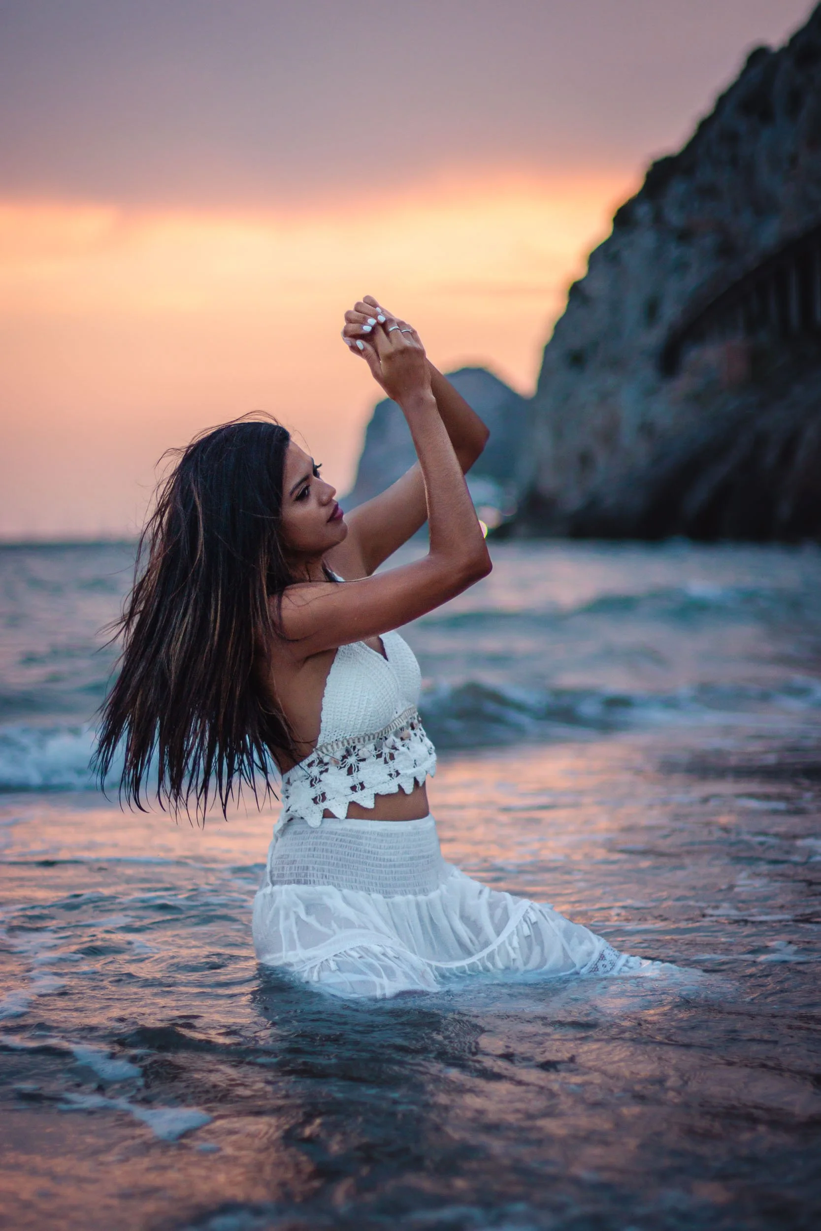 Side view of a woman standing in shallow water and lifting her arms at sunset