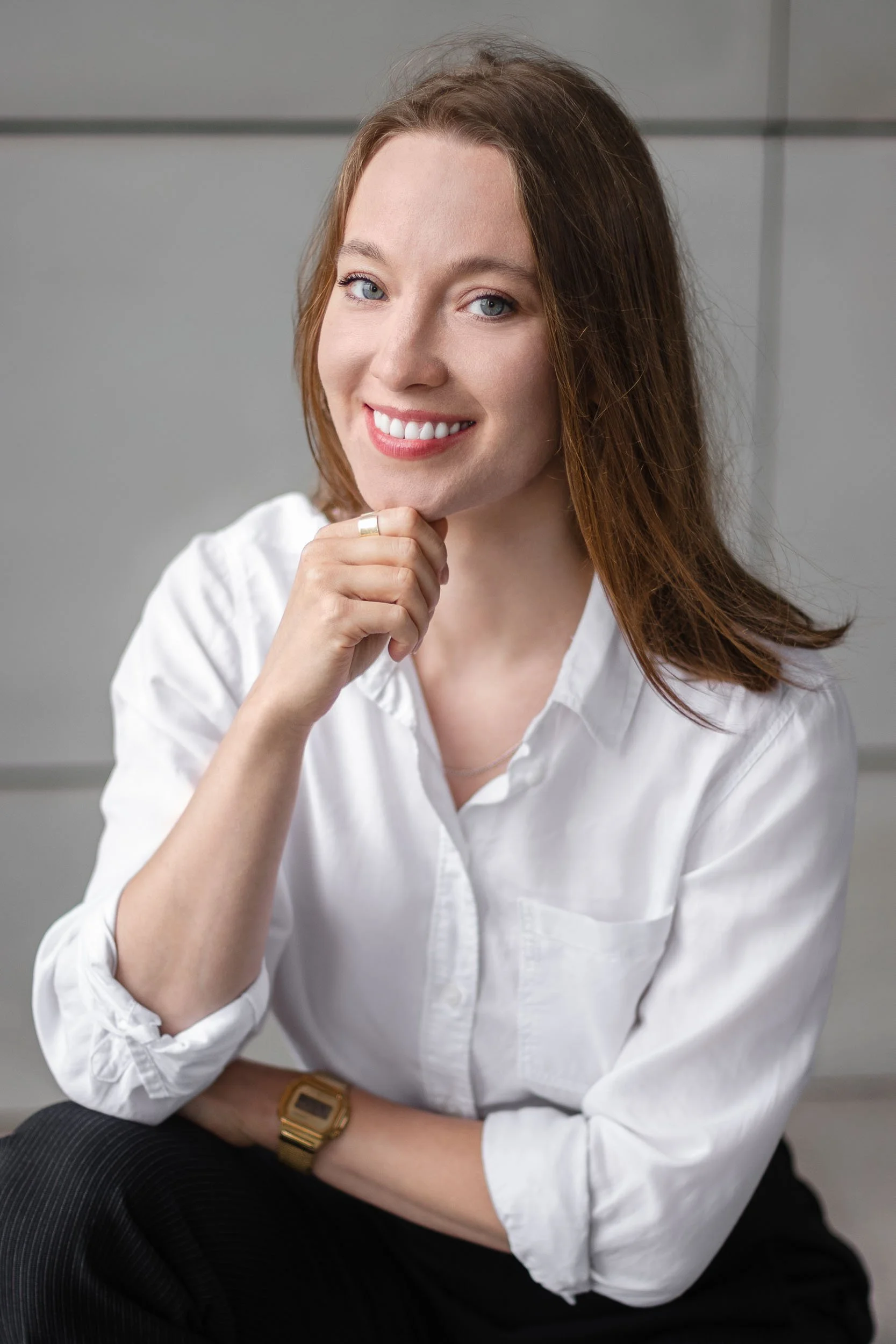 Headshot of a woman smiling to the camera wearing a white shirt and posing with her hand around her chin in front of a grey wall.