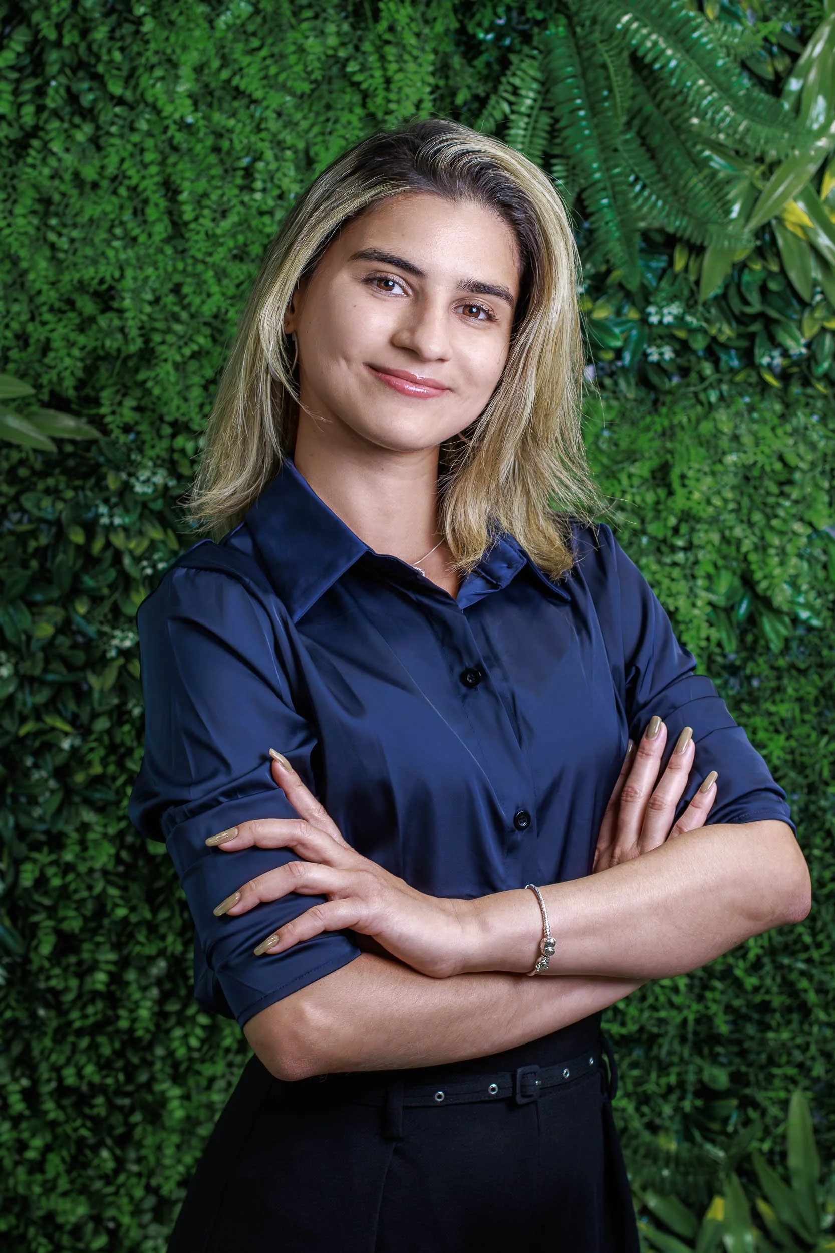 Professional headshot of a woman in a navy shirt standing with arms crossed against a green plant wall