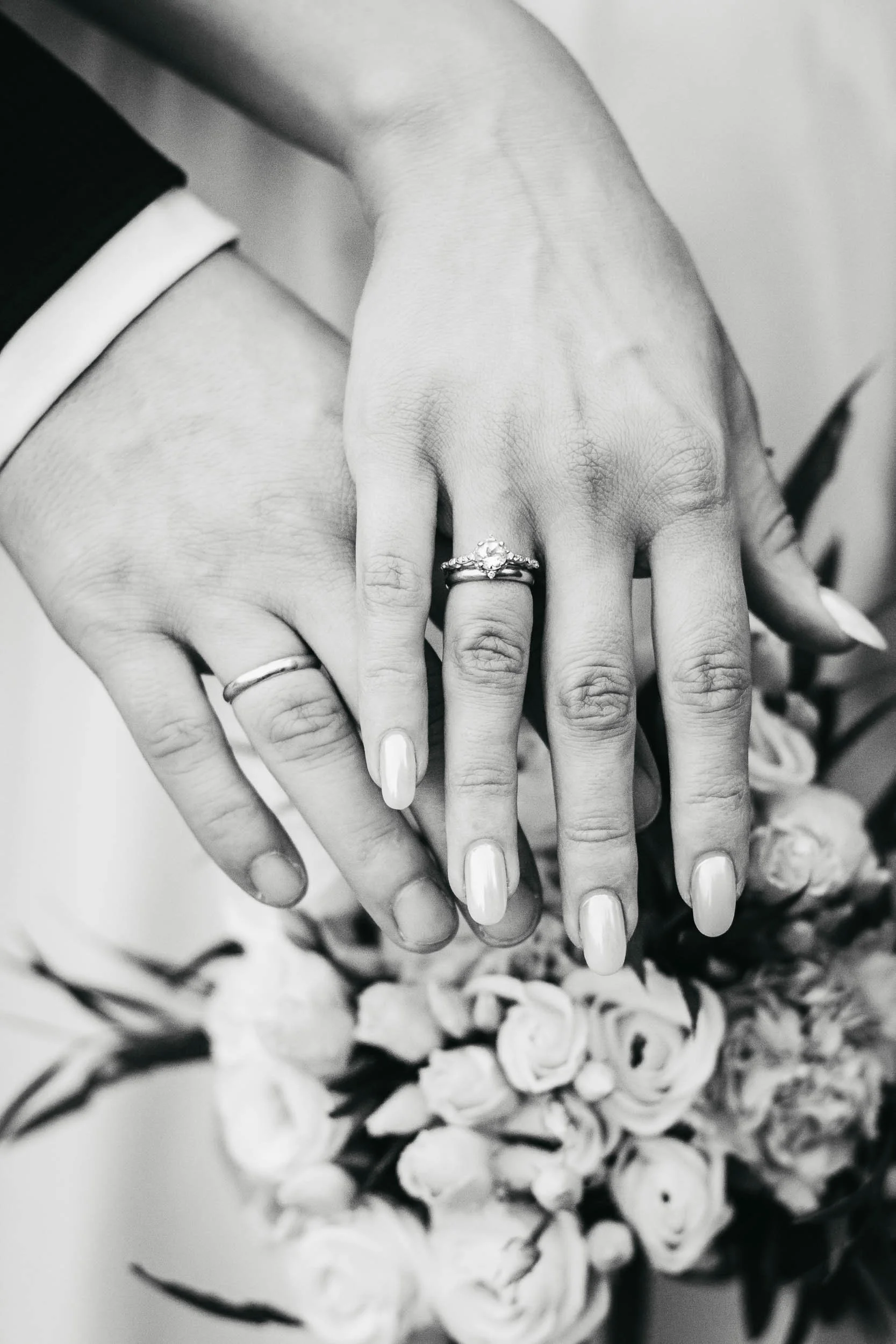 Black and white close-up of the couple's hands with wedding rings resting over the bouquet