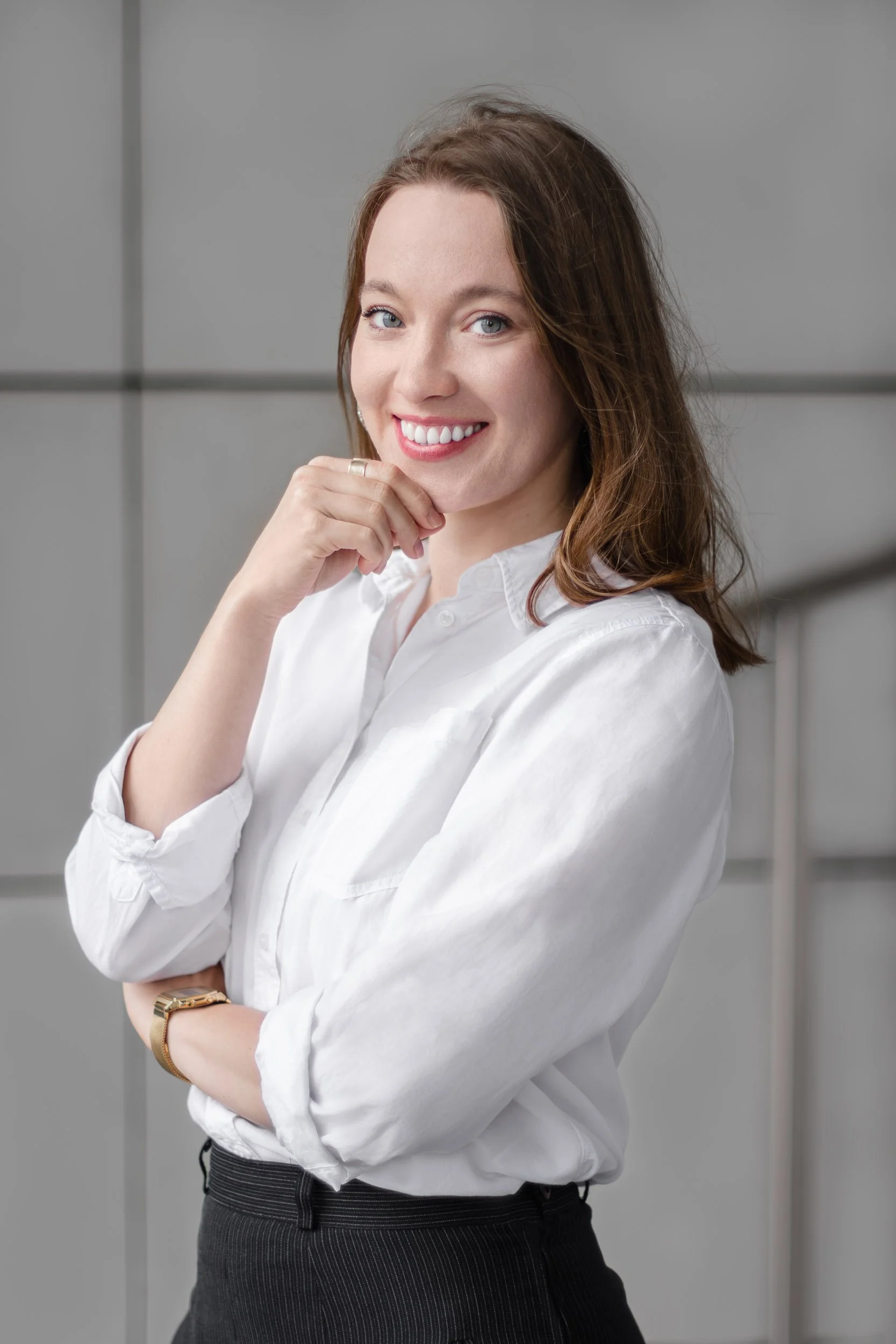 Professional headshot of a smiling woman in a white shirt posing with one hand near her chin against a grey wall