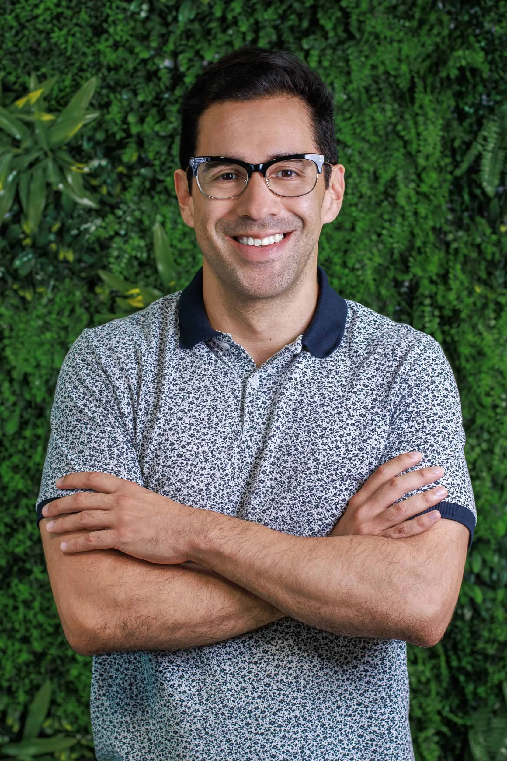 Professional headshot of a man wearing glasses standing with arms crossed against a green plant wall