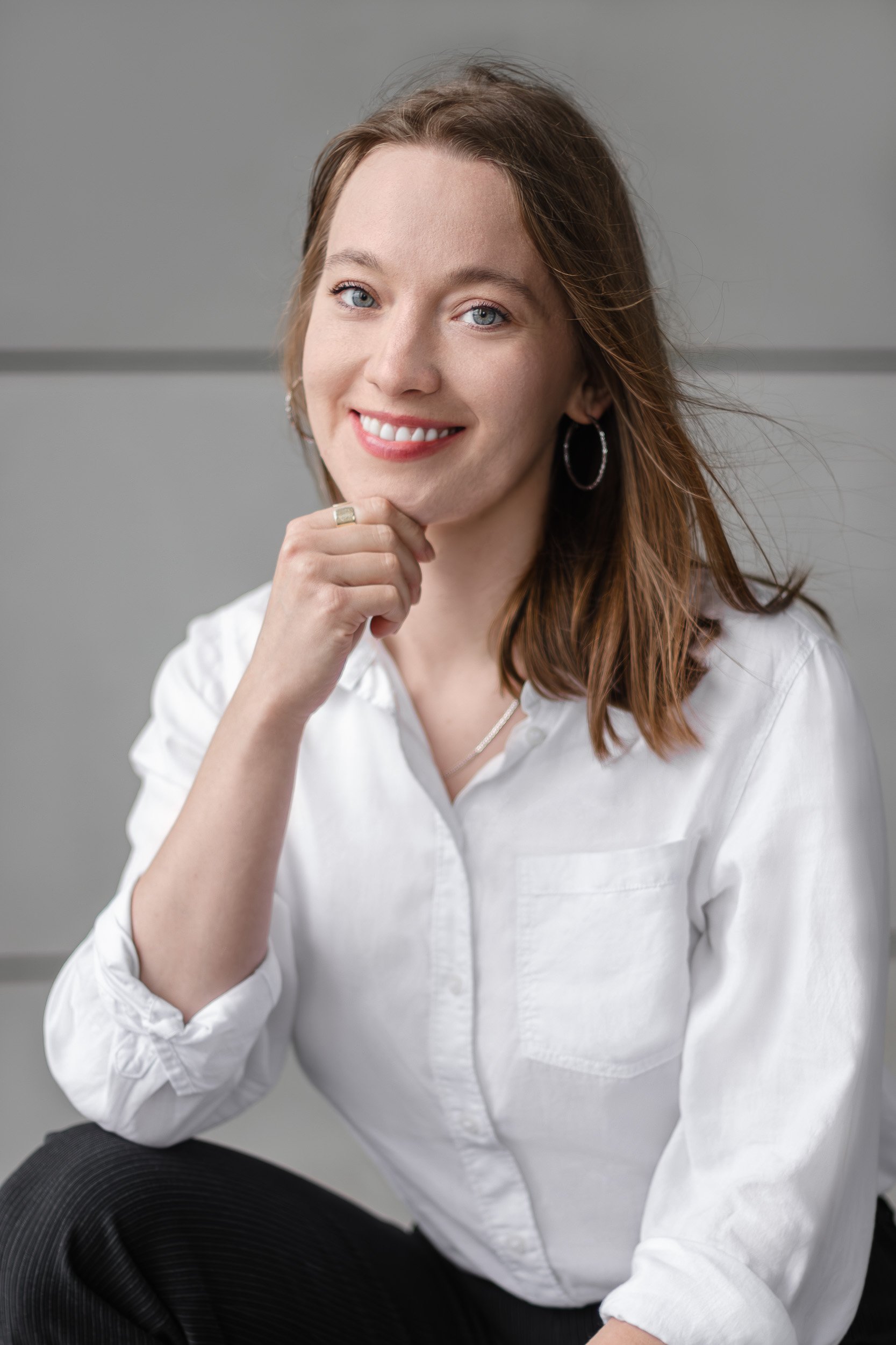 Professional headshot of a smiling woman in a white shirt posing with one hand near her chin against a grey wall