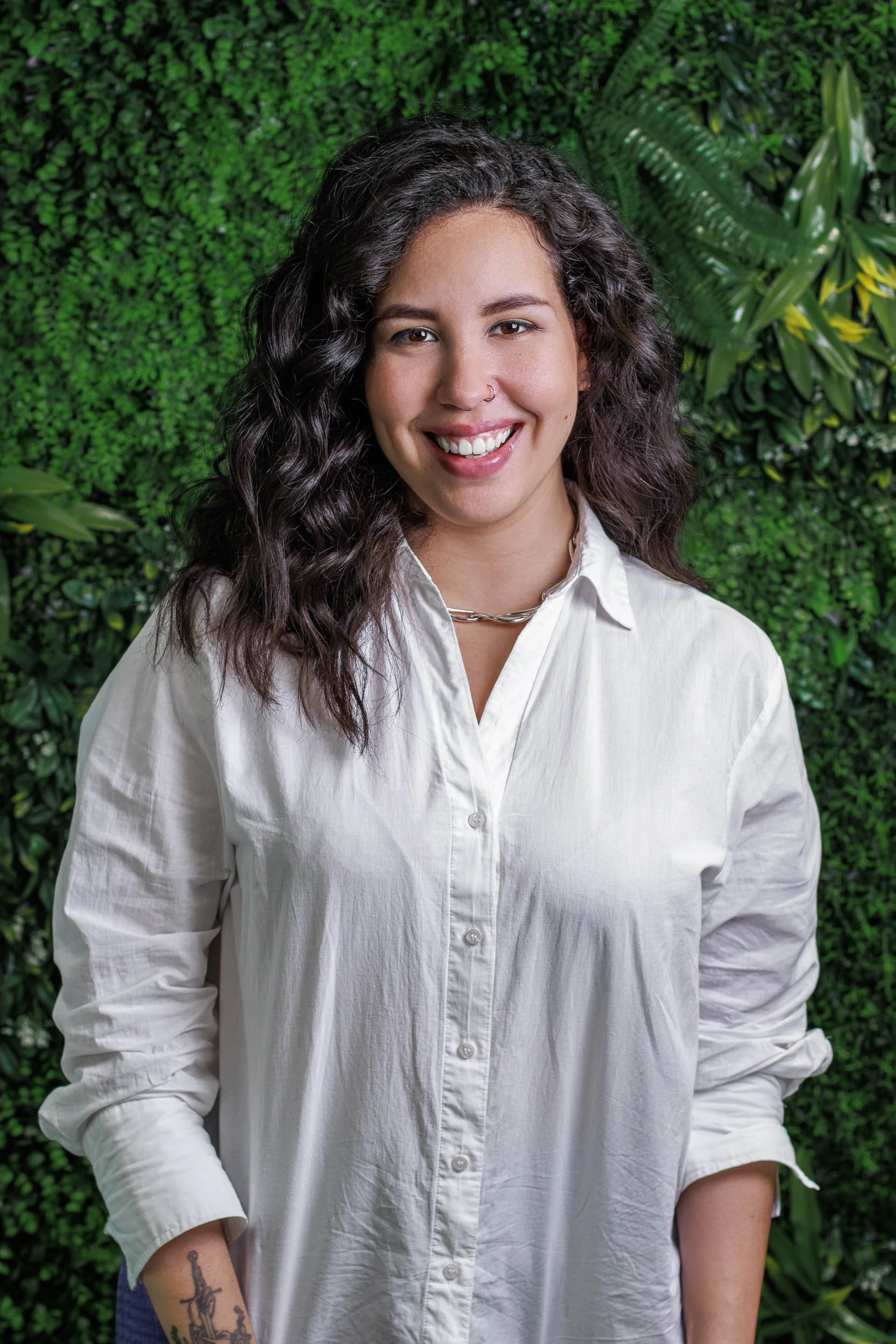 Professional headshot of a smiling woman in a white shirt standing against a green plant wall