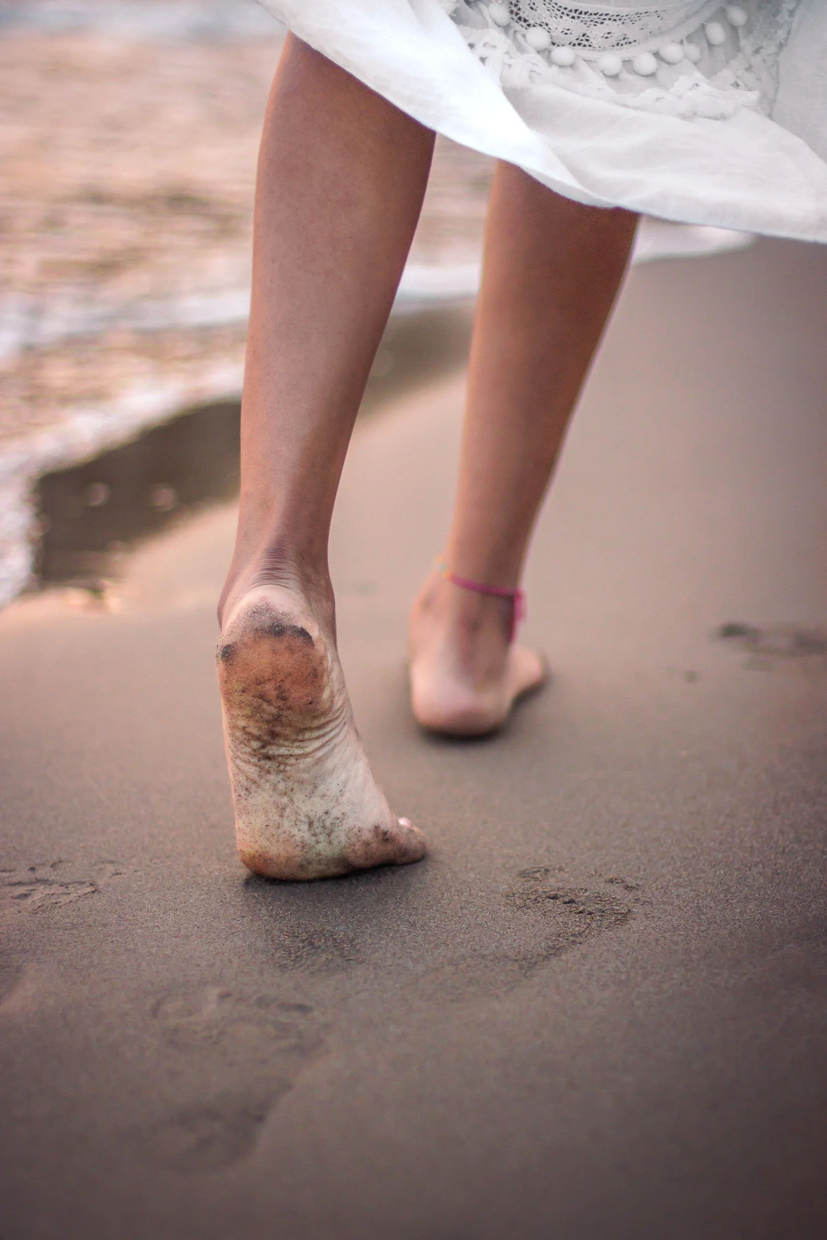 Close-up detail of bare feet and the hem of a white dress on wet sand