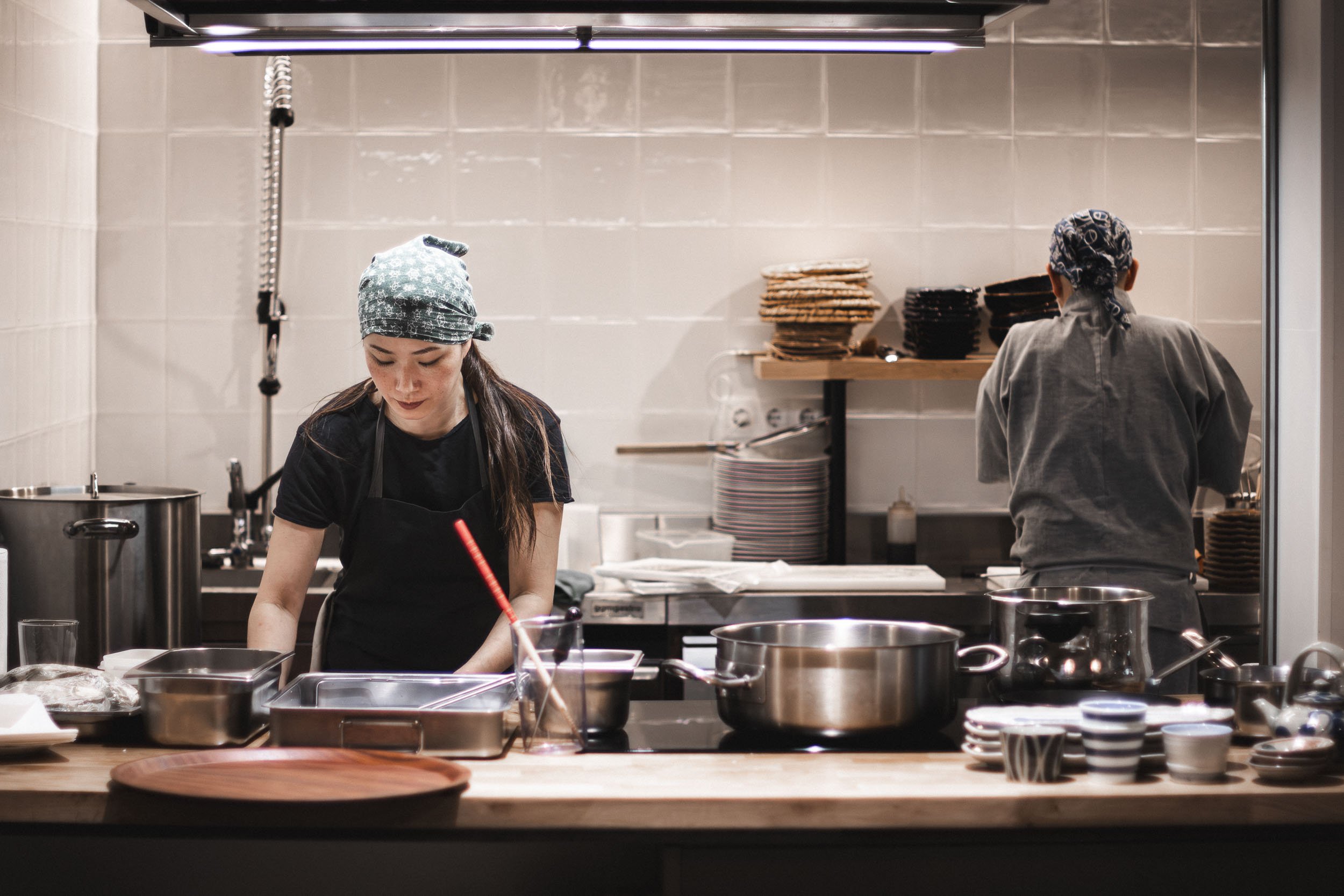Two chefs working in an open restaurant kitchen, with one standing at the counter in the foreground and another preparing food in the background.
