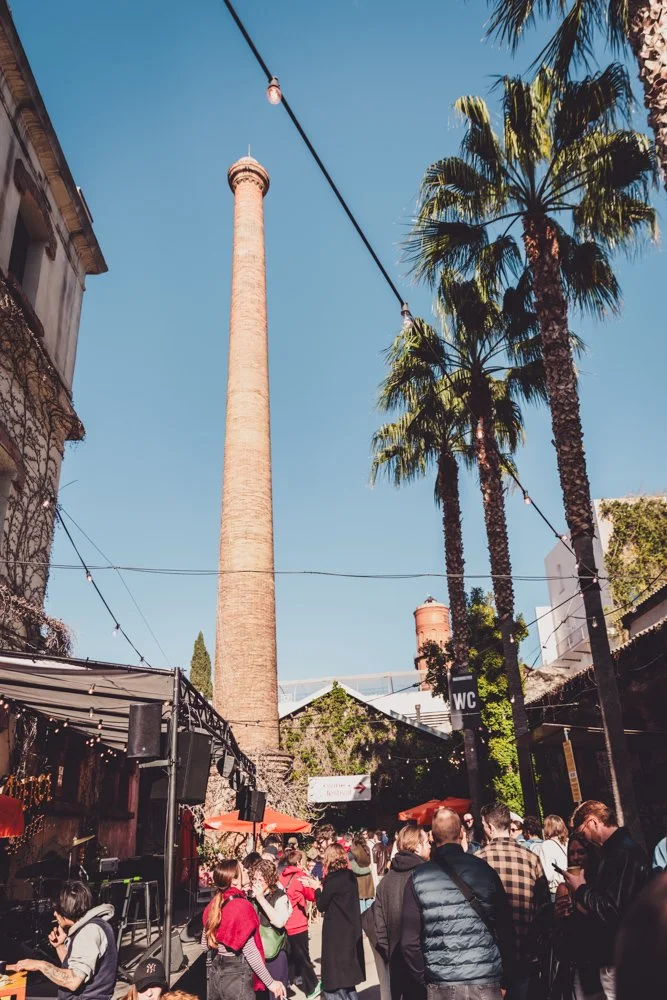 Exterior view of an event venue with a tall chimney, palm trees, and people walking below