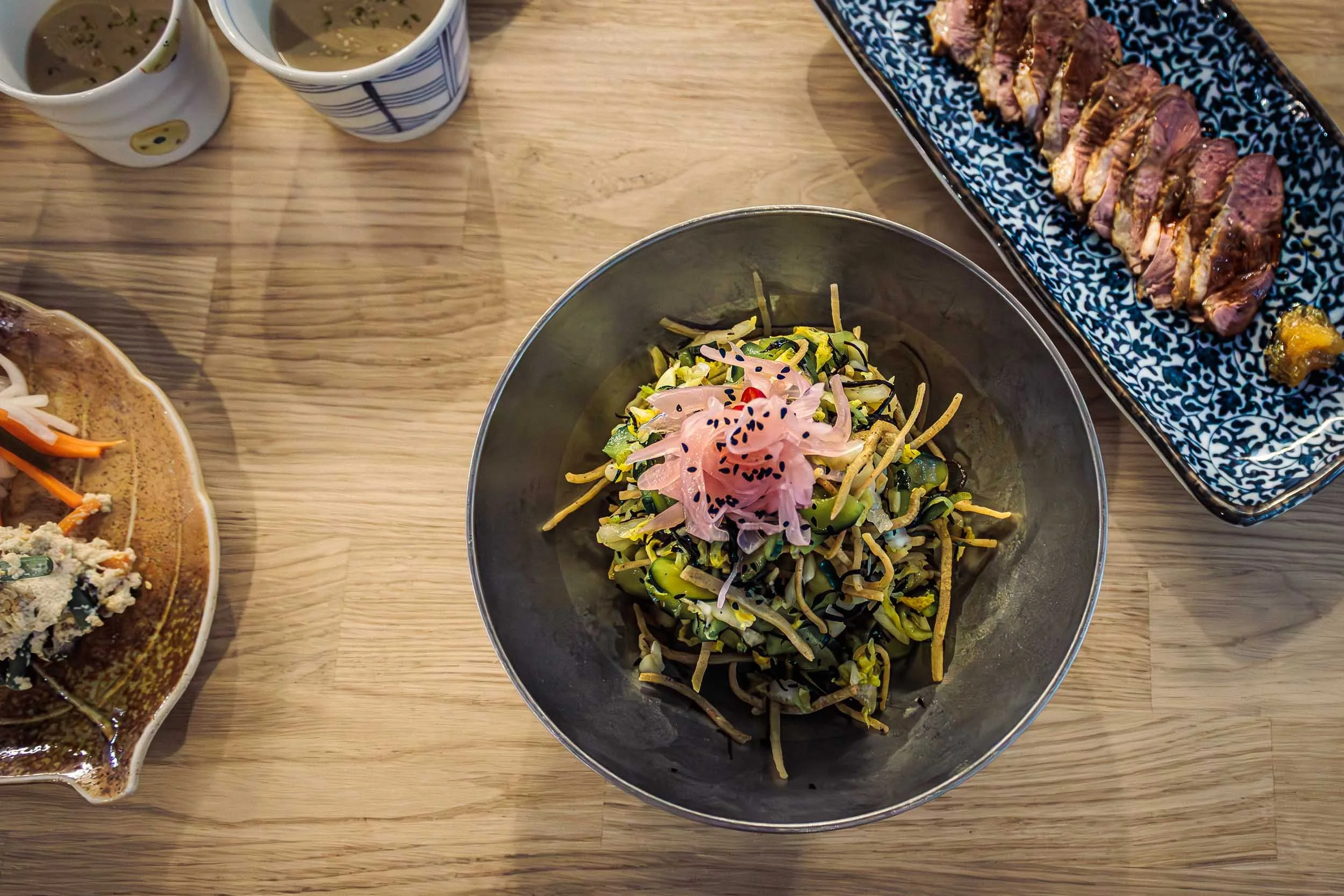 Overhead view of plated dishes on a wooden table, with a bowl of food in the center and other plates partially visible around it.